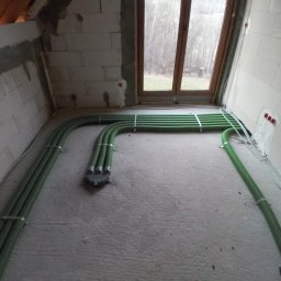 Grzegorz Dudzik - Zielone rury wentylacyjne prowadzone po podłodze surowego pomieszczenia z oknem, widoczne mocowania do ściany z betonu komórkowego.