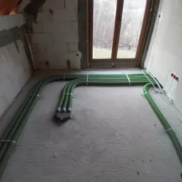 Zielone rury wentylacyjne prowadzone po podłodze surowego pomieszczenia z oknem, widoczne mocowania do ściany z betonu komórkowego.