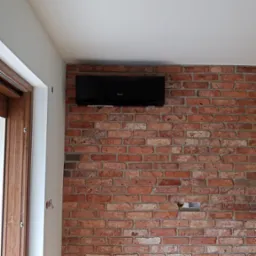 Czarna jednostka klimatyzacyjna zamontowana na ścianie z czerwonej cegły w nowoczesnym wnętrzu z drewnianą podłogą i białymi ścianami.