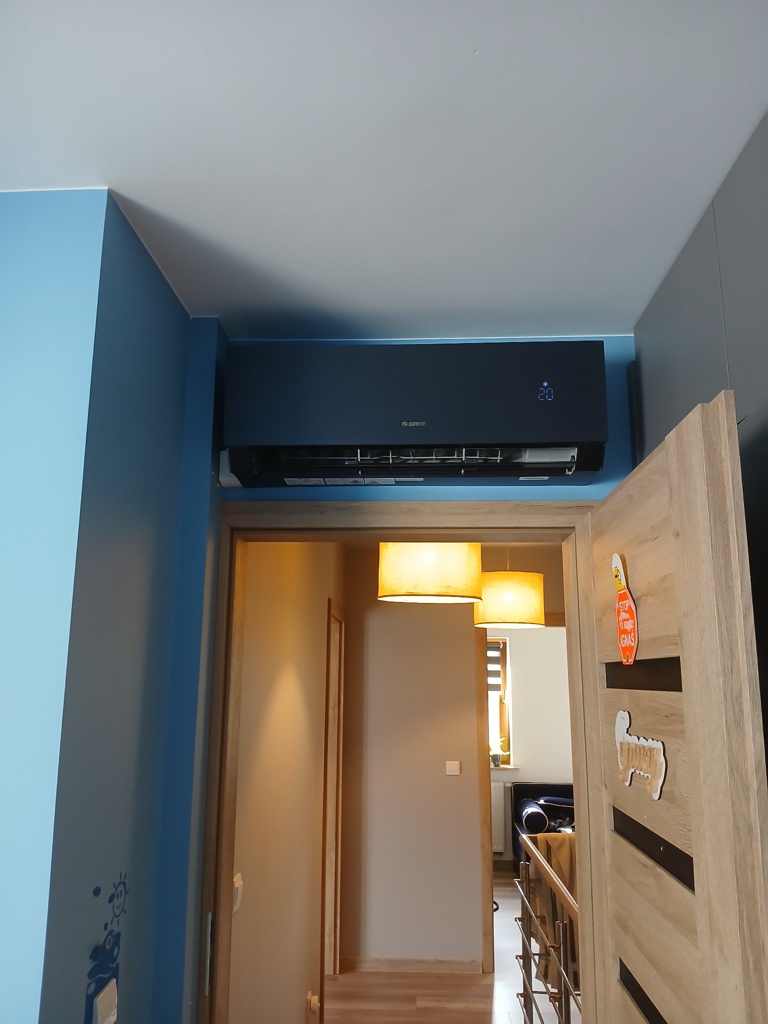 Czarna jednostka klimatyzacji zamontowana na ścianie w pokoju dziecięcym, widoczna nad otwartymi drzwiami, z dekoracjami na drzwiach i niebieską ścianą po lewej stronie.