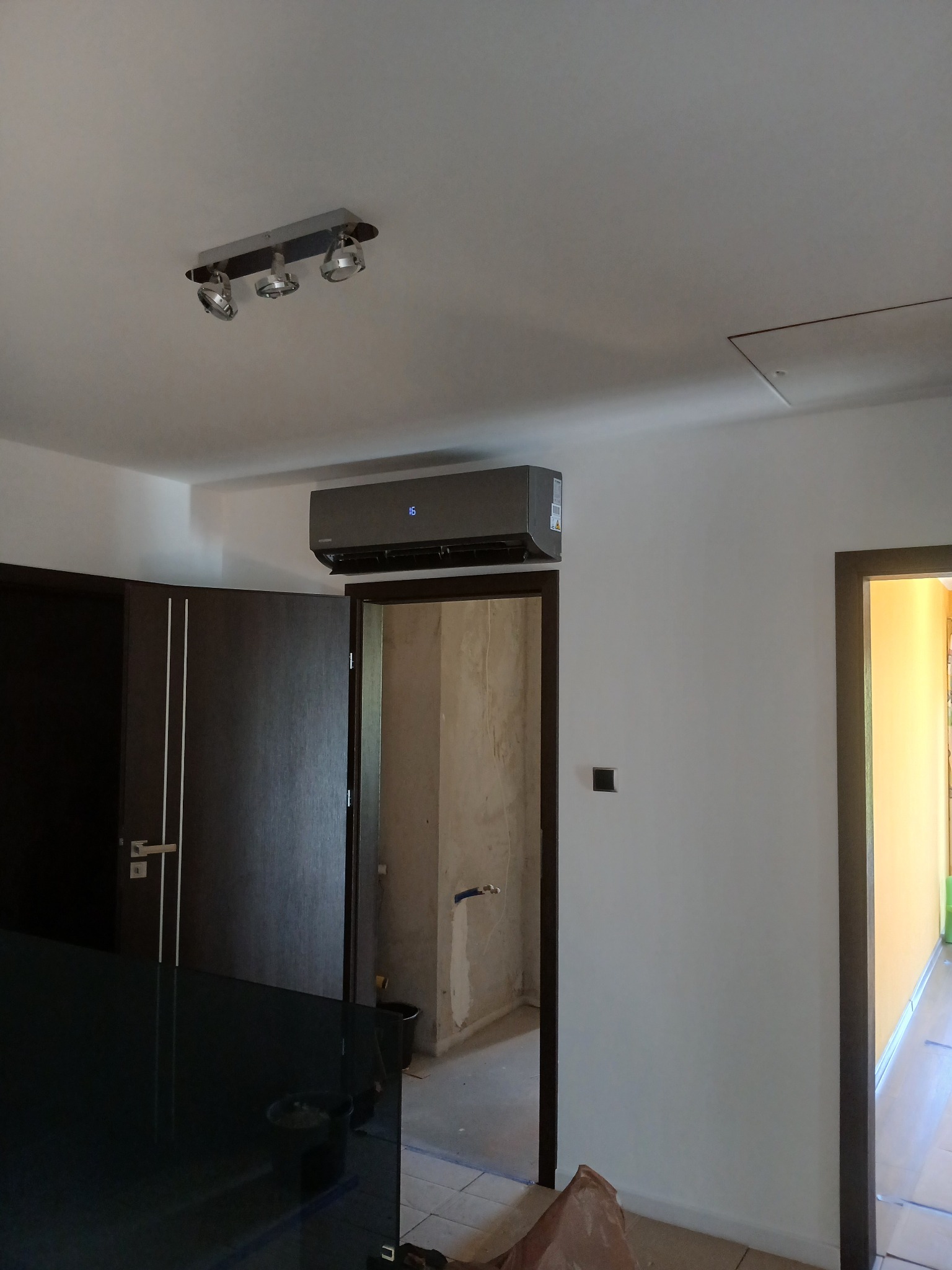 Szary klimatyzator ścienny z wyświetlaczem temperatury, zamontowany w pomieszczeniu z widocznymi drzwiami i elementami wykończeniowymi.