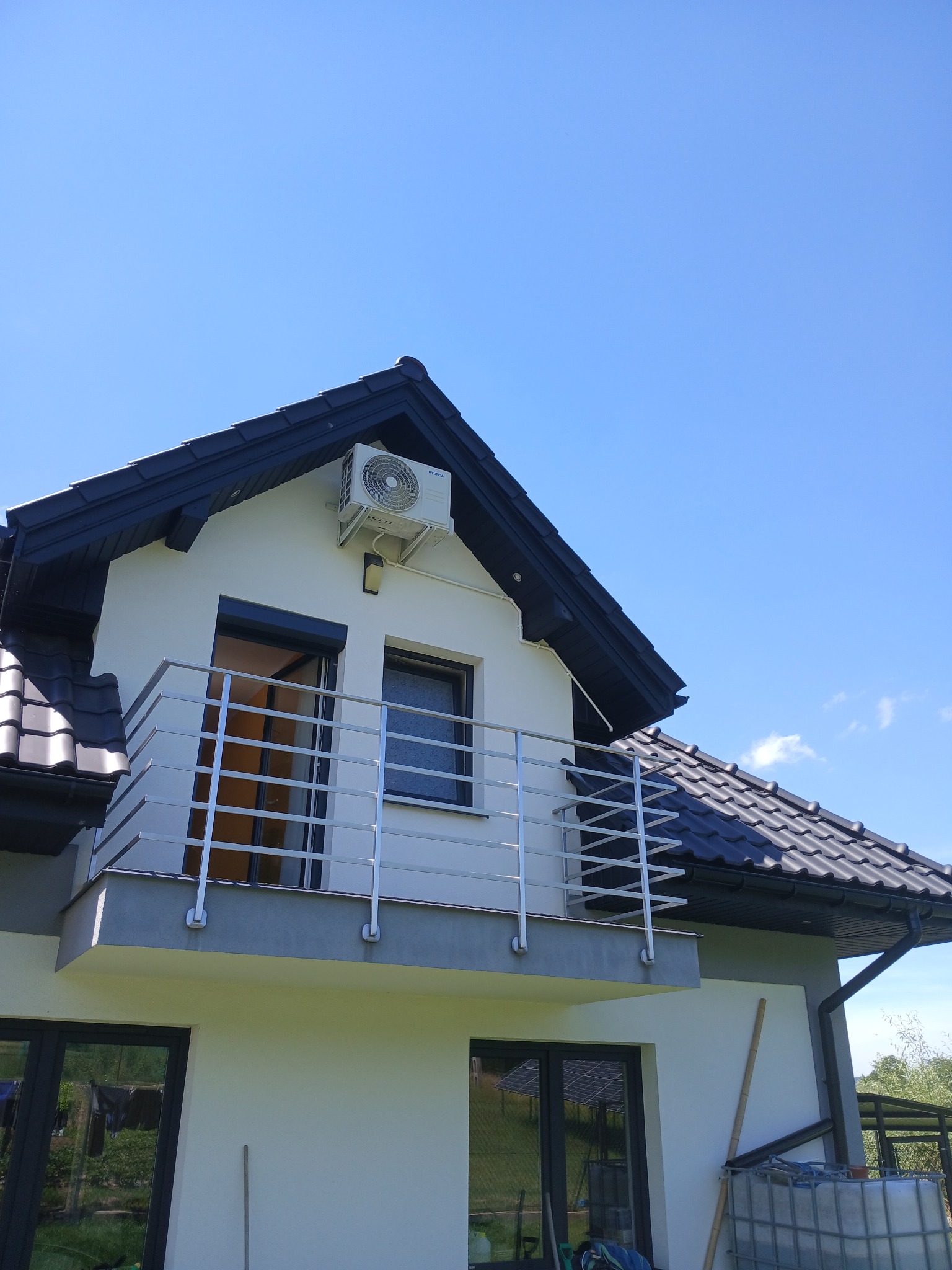 Biały klimatyzator zamontowany na elewacji domu z balkonem, widok z dołu na błękitne niebo.