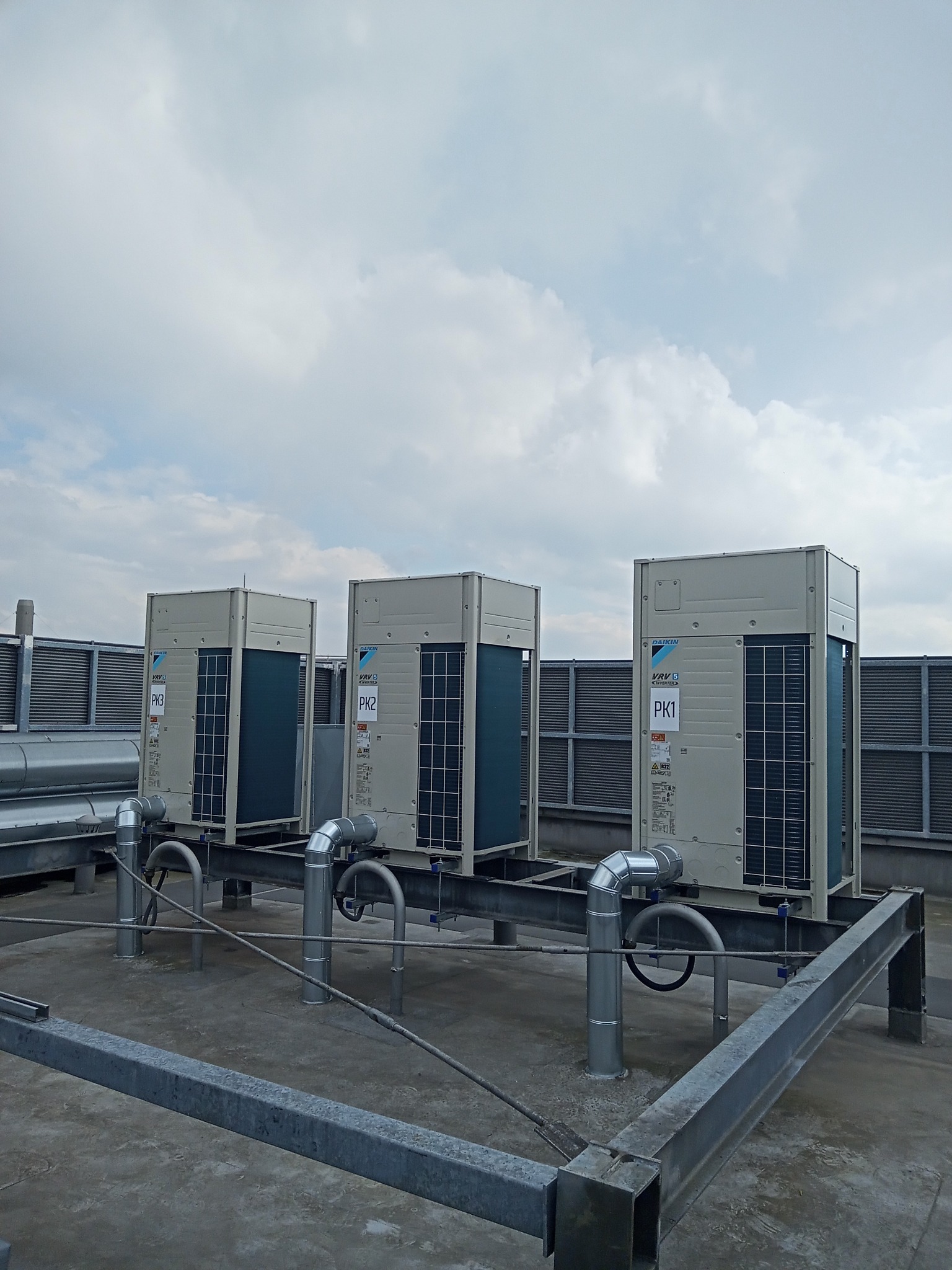 Trzy jednostki zewnętrzne klimatyzacji Daikin VRV 6 Inverter, oznaczone PK1, PK2 i PK3, zainstalowane na dachu budynku, z widocznymi rurami i metalową konstrukcją wsporczą, na tle pochmurnego nieba.