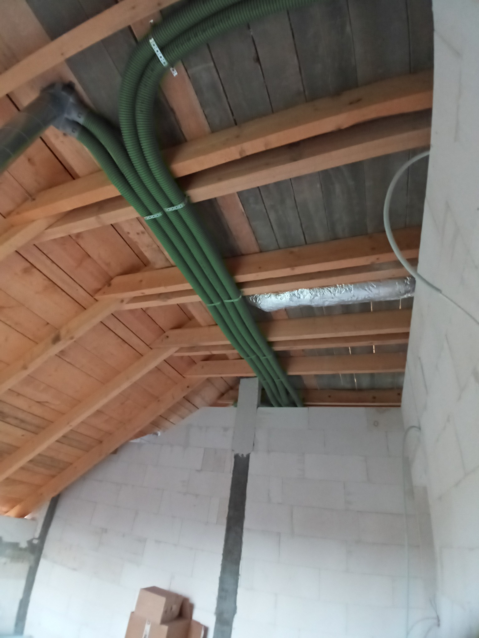 Zielone rury wentylacyjne przymocowane do drewnianej konstrukcji dachu na poddaszu, z widoczną izolacją termiczną i fragmentem ściany z bloczków betonowych.