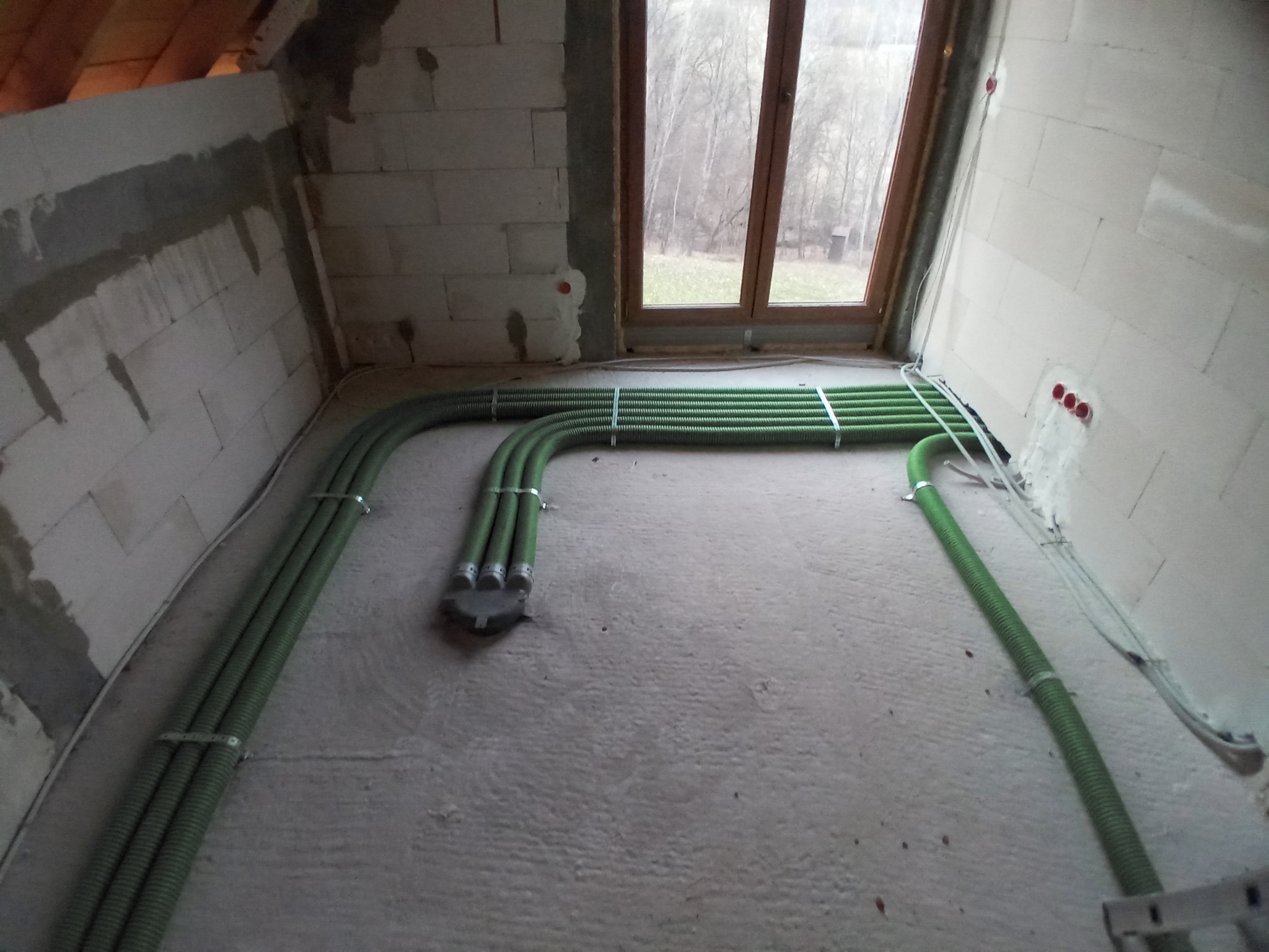 Zielone rury wentylacyjne prowadzone po podłodze surowego pomieszczenia z oknem, widoczne mocowania do ściany z betonu komórkowego.