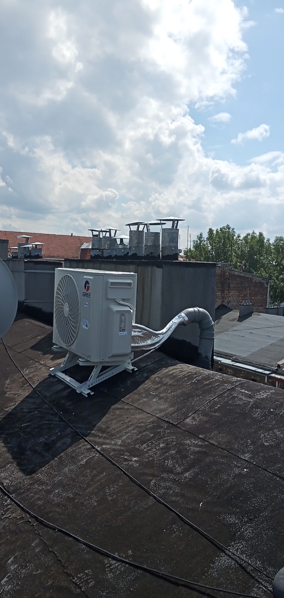 Jednostka zewnętrzna klimatyzatora marki Gree zamontowana na metalowym stelażu na płaskim, czarnym dachu, z widocznymi przewodami w izolacji aluminiowej, w tle kominy i budynki pod błękitnym niebem...