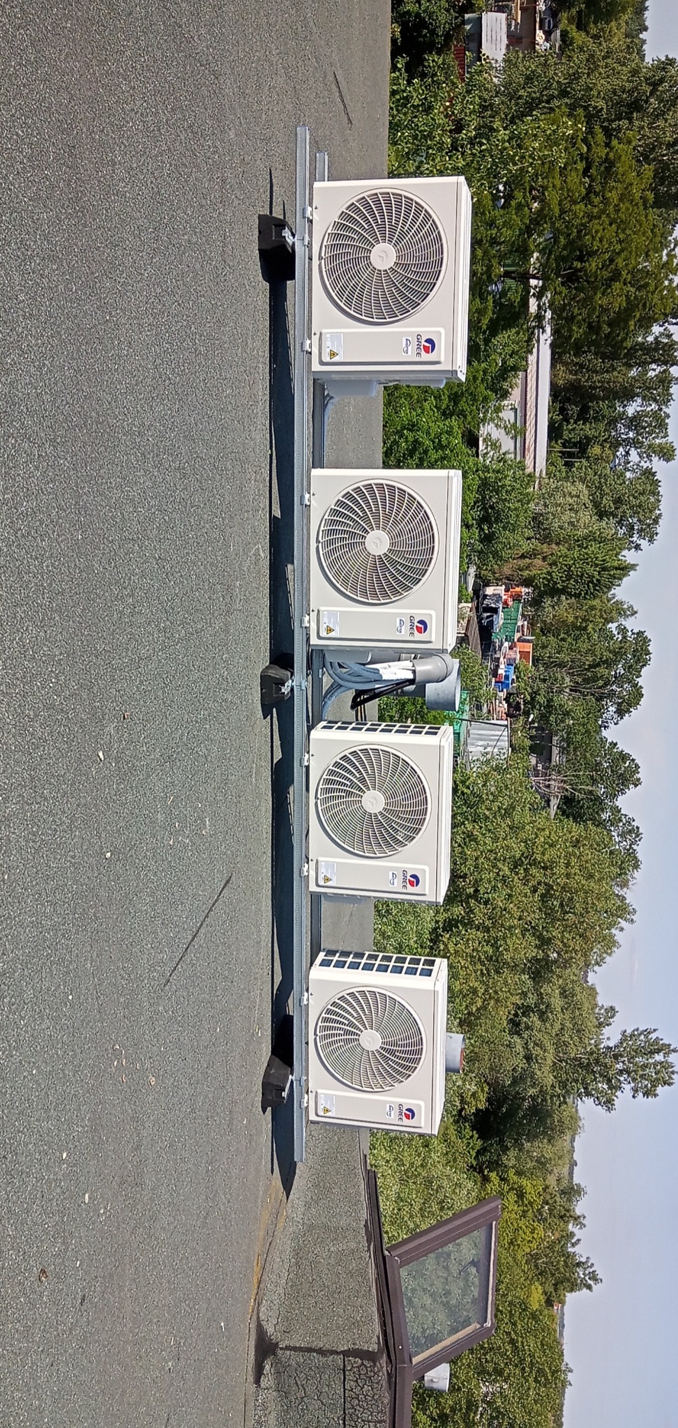 Cztery jednostki zewnętrzne klimatyzacji marki Gree zamontowane na dachu budynku, widoczne elementy instalacji i otaczająca zieleń.