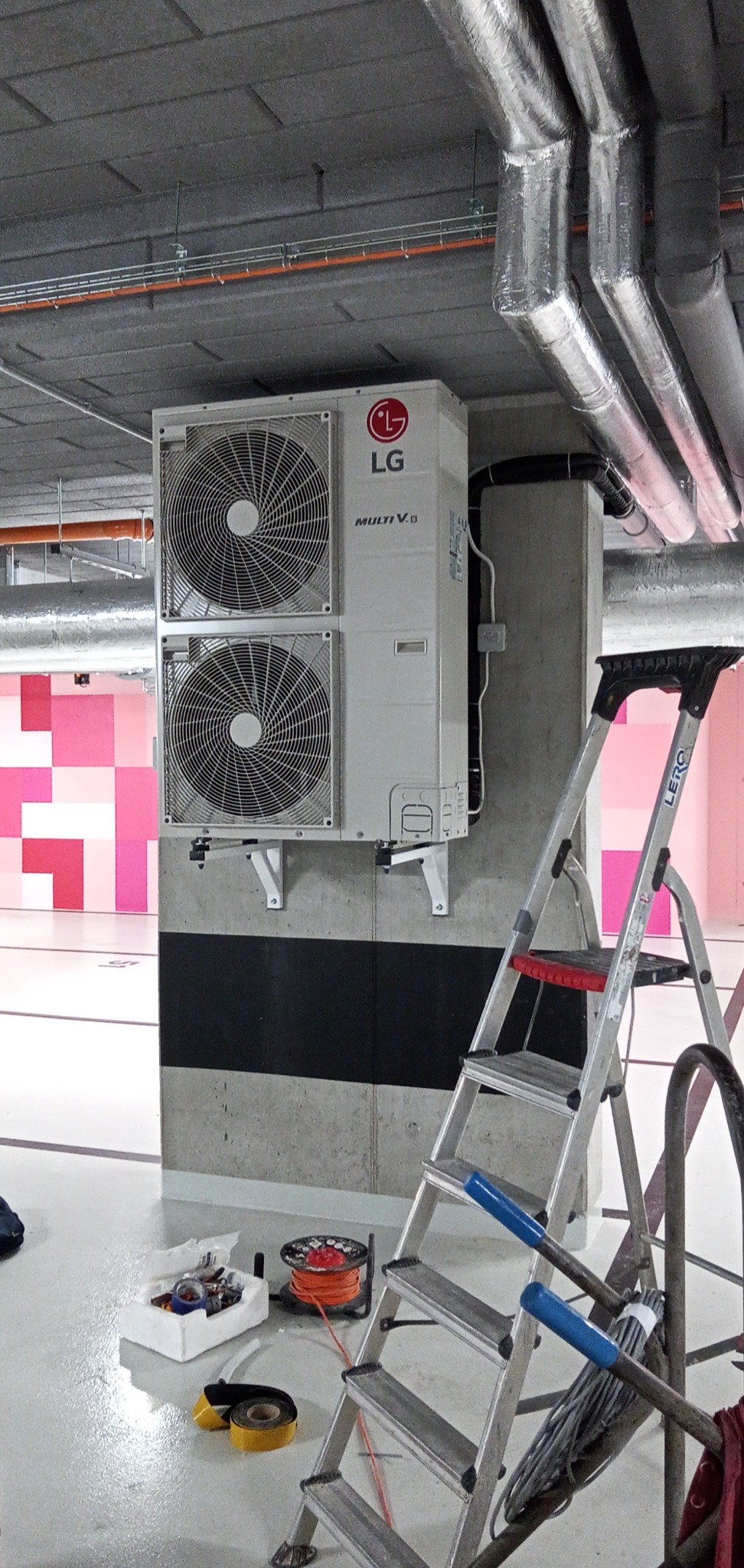 Dwu-wentylatorowa jednostka zewnętrzna LG Multi V zamontowana na betonowym słupie w garażu podziemnym, widoczna drabina i narzędzia instalacyjne.