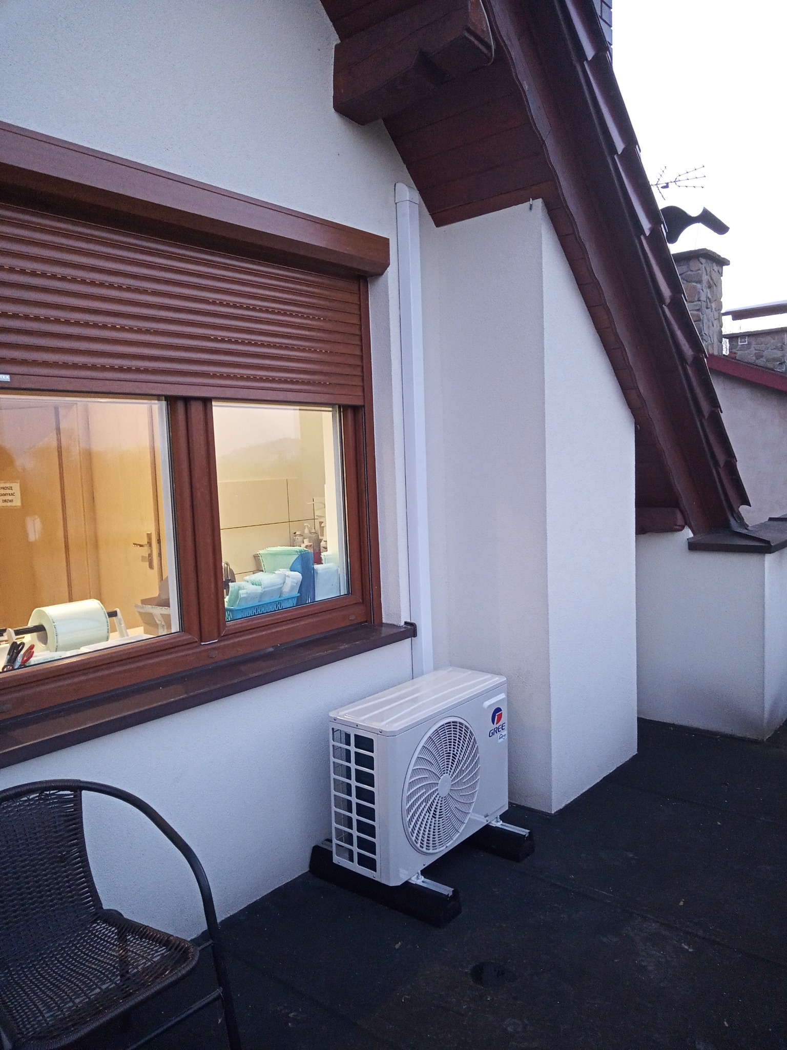 Zewnętrzna jednostka klimatyzatora marki Gree zamontowana na czarnym dachu budynku z brązowymi okiennicami, widoczny fragment krzesła z plecionki.