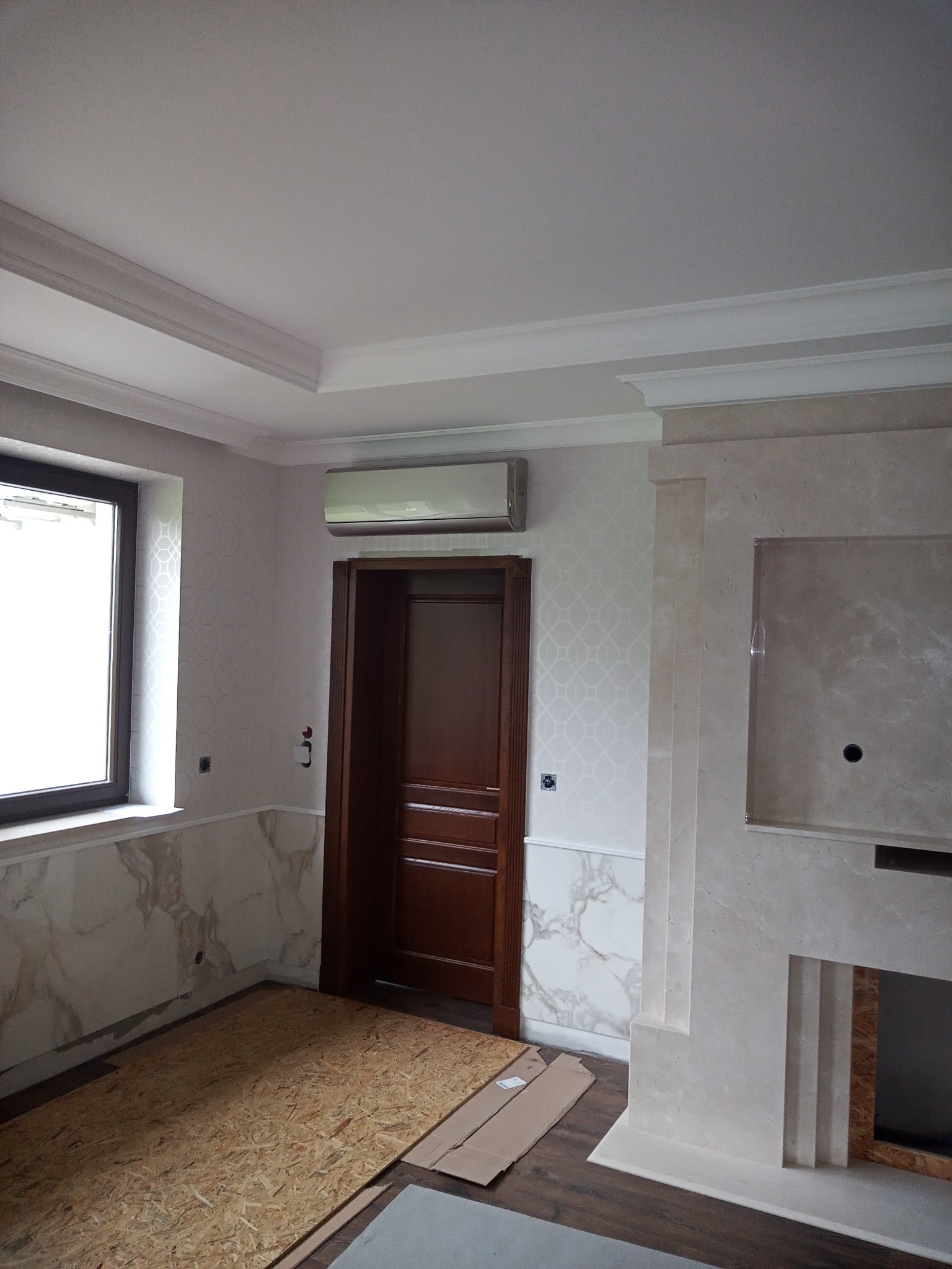 Klimatyzator ścienny zamontowany w pomieszczeniu w Raciechowicach, widoczny na tle wzorzystej tapety i drewnianej stolarki drzwiowej, z fragmentem marmurowego obramowania kominka po prawej stronie.