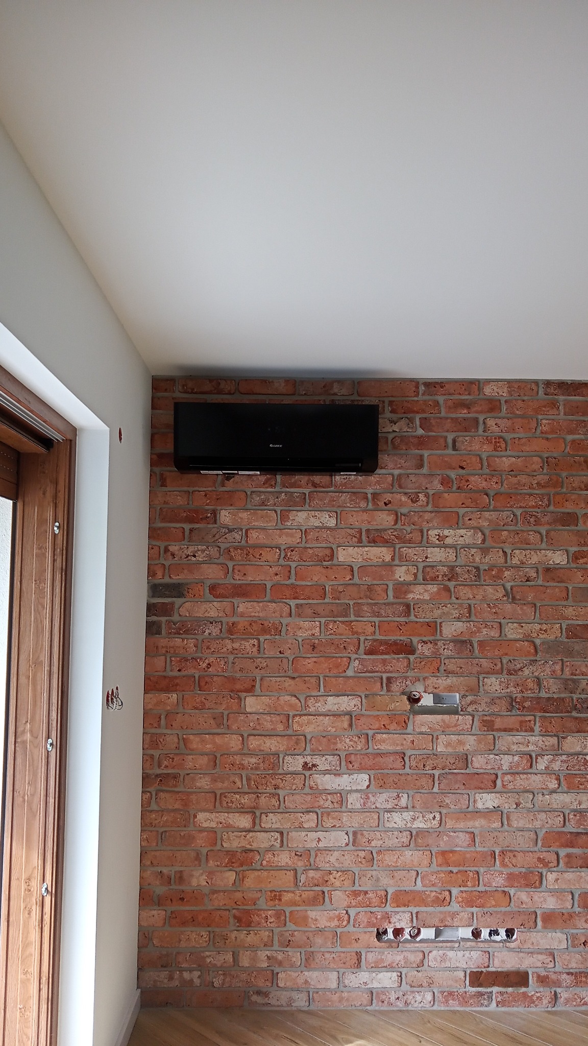 Czarna jednostka klimatyzacyjna zamontowana na ścianie z czerwonej cegły w nowoczesnym wnętrzu z drewnianą podłogą i białymi ścianami.