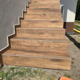 Zewnętrzne schody wejściowe wyłożone brązowymi płytkami imitującymi drewno, z białymi listwami wykończeniowymi, prowadzące do budynku z elewacją w dwóch odcieniach szarości.