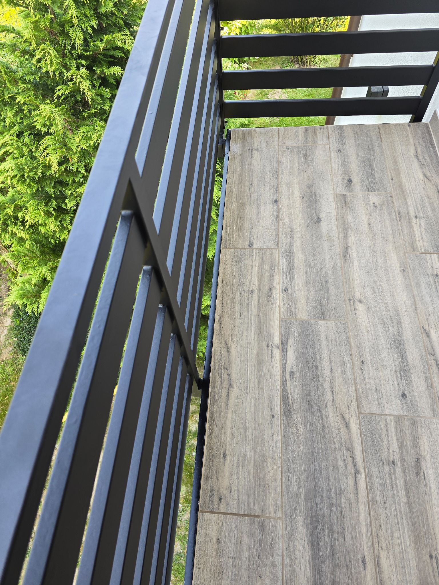 Wykończony balkon z szarą podłogą imitującą drewno i nowoczesną, czarną balustradą na tle zieleni.