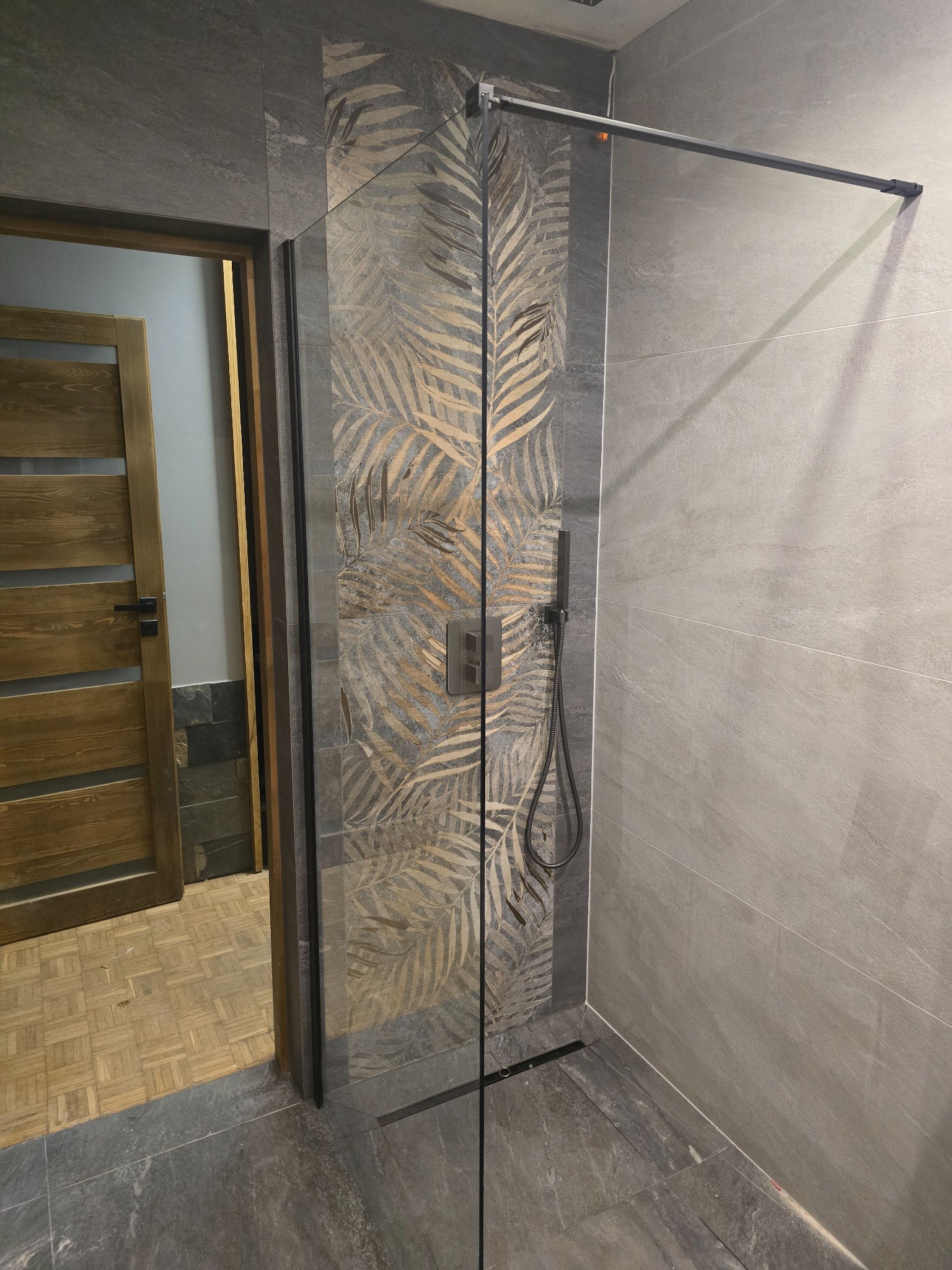 Szklana kabina prysznicowa z czarnymi profilami, przylegająca do ściany wyłożonej szarą płytką z motywem złotych liści palmowych, widoczna drewniana podłoga i drzwi w tle.