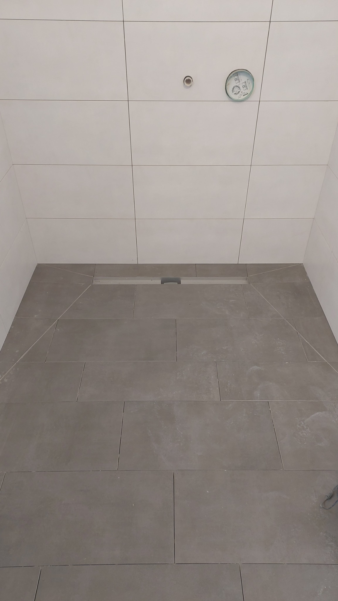 Nowoczesna kabina prysznicowa z szarymi płytkami podłogowymi i białymi płytkami ściennymi, widoczny odpływ liniowy i instalacja pod natrysk.