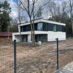 SILVER SUMMIT DEVELOPMENT SPÓŁKA Z OGRANICZONĄ ODPOWIEDZIALNOŚCIĄ - Nowoczesny, dwukondygnacyjny dom z płaskim dachem i dużymi oknami, otoczony drzewami i ogrodzeniem z siatki. Widoczne przygotowane podłoże wokół budynku.