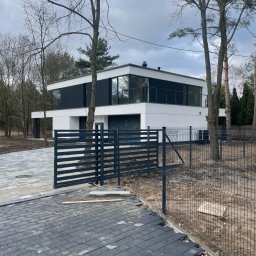 SILVER SUMMIT DEVELOPMENT SPÓŁKA Z OGRANICZONĄ ODPOWIEDZIALNOŚCIĄ - Nowoczesny, dwukondygnacyjny dom z płaskim dachem, czarnymi akcentami i dużymi oknami, widziany zza czarnego ogrodzenia panelowego na nierównym podjeździe z kostki brukowej i ziemi.