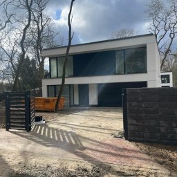 SILVER SUMMIT DEVELOPMENT SPÓŁKA Z OGRANICZONĄ ODPOWIEDZIALNOŚCIĄ - Nowoczesny, dwukondygnacyjny dom w trakcie budowy, z dużymi oknami i płaskim dachem, widoczny pomarańczowy kontener na gruz i fragment ogrodzenia.