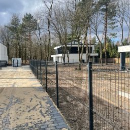 SILVER SUMMIT DEVELOPMENT SPÓŁKA Z OGRANICZONĄ ODPOWIEDZIALNOŚCIĄ - Nowoczesny dom z płaskim dachem i dużymi oknami, widoczny zza nowego, metalowego ogrodzenia panelowego, otoczony drzewami bez liści w pochmurny dzień.