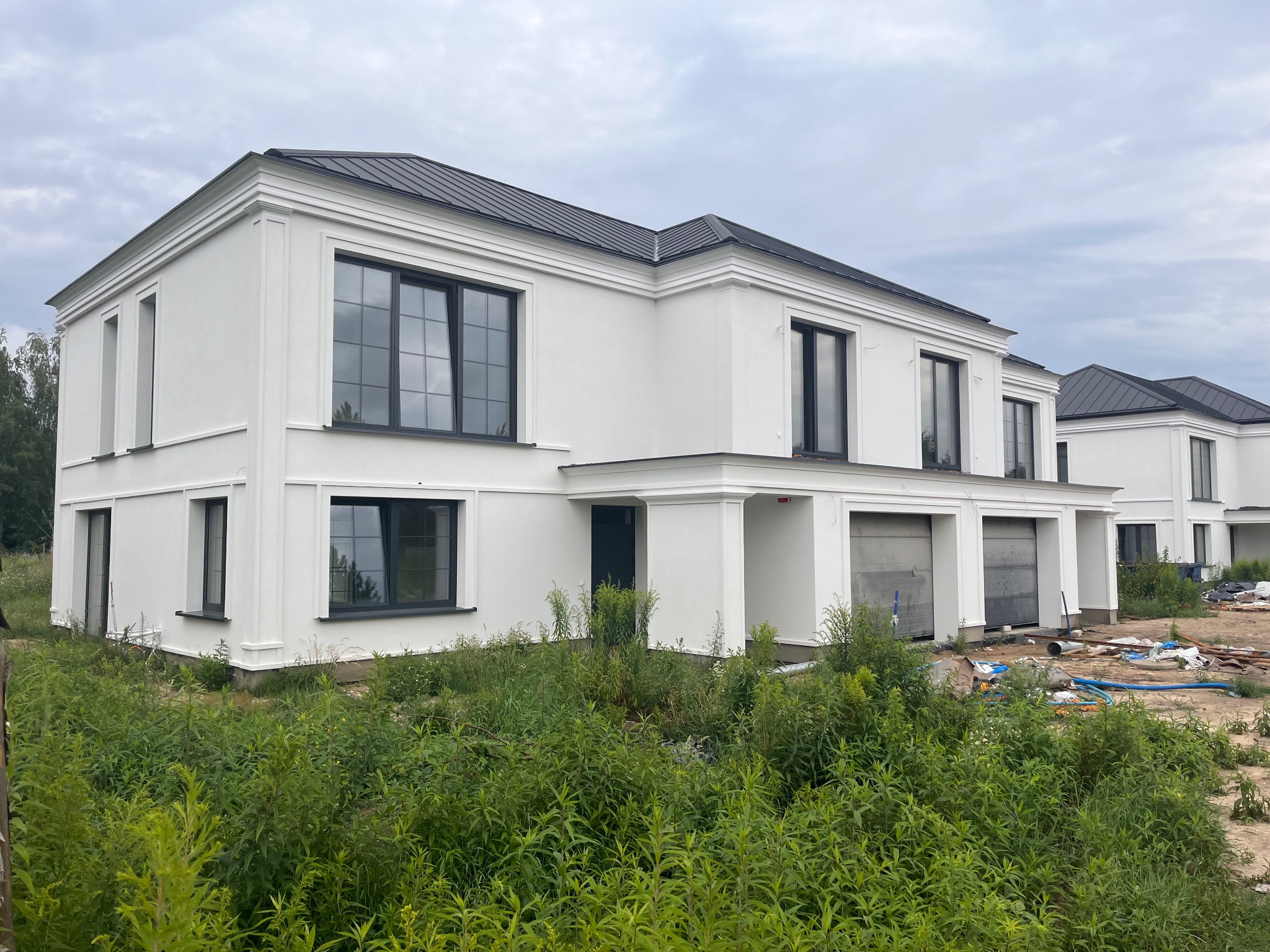 Nowoczesny, dwukondygnacyjny biały budynek z czarnym dachem i oknami, w surowym stanie, widoczny od frontu, otoczony wysoką trawą i elementami placu budowy.