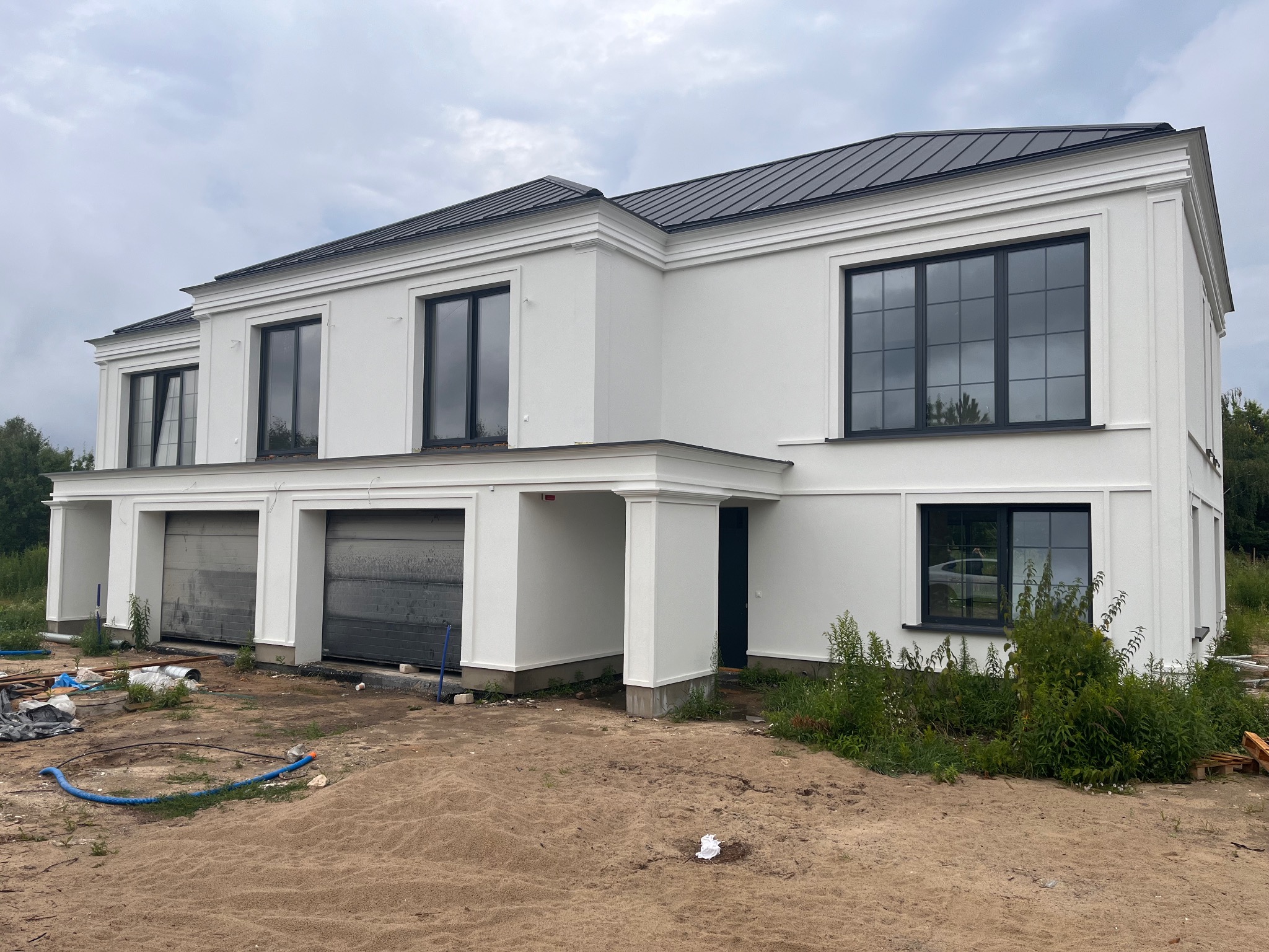 Dwukondygnacyjny budynek w stanie surowym zamkniętym z garażem dwustanowiskowym, biała elewacja, czarne okna i dach, teren wokół w trakcie prac wykończeniowych.