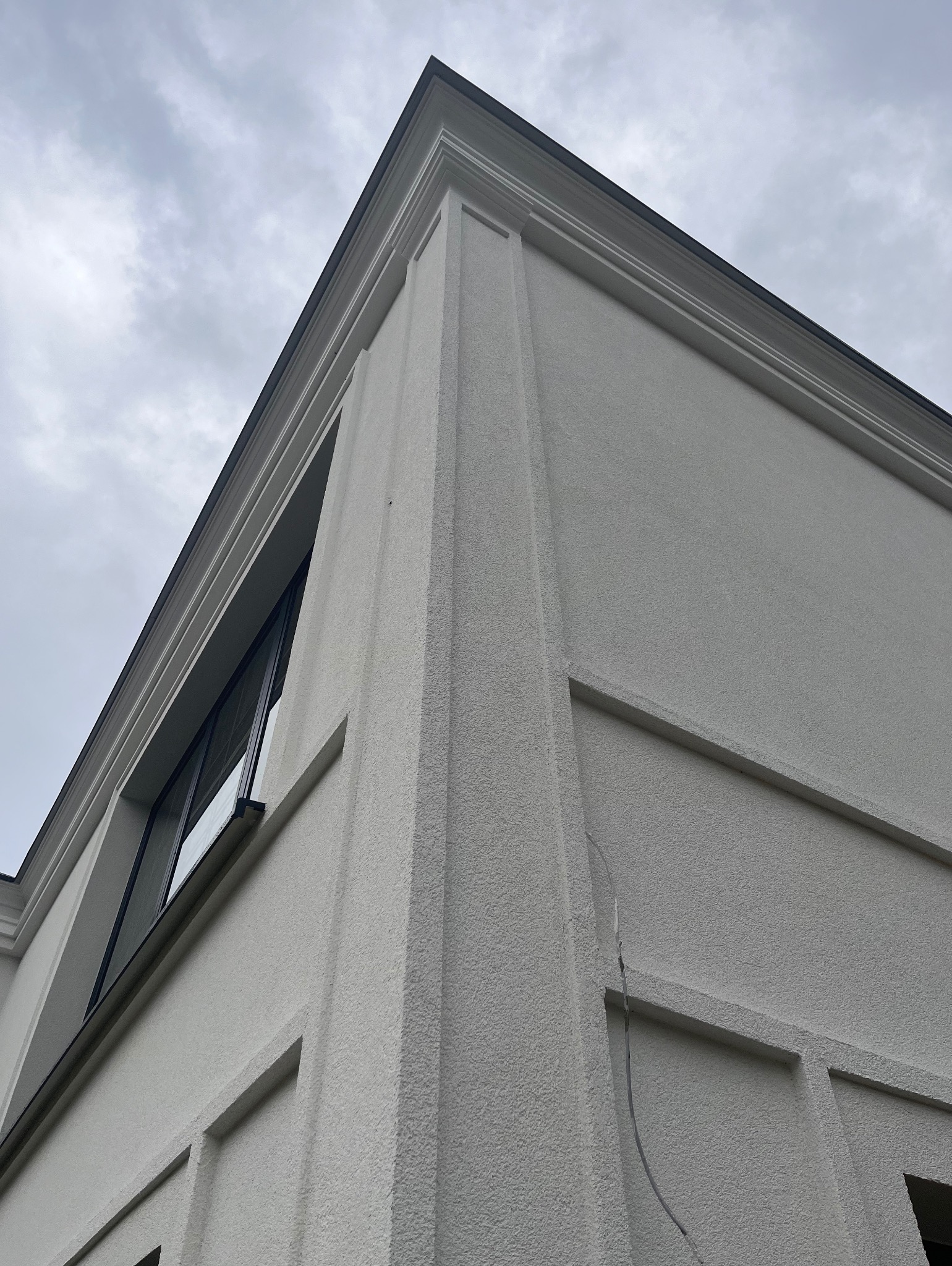 Fragment narożnika budynku z jasną elewacją o strukturze baranka, widoczne okno z czarną ramą i dekoracyjne elementy architektoniczne.