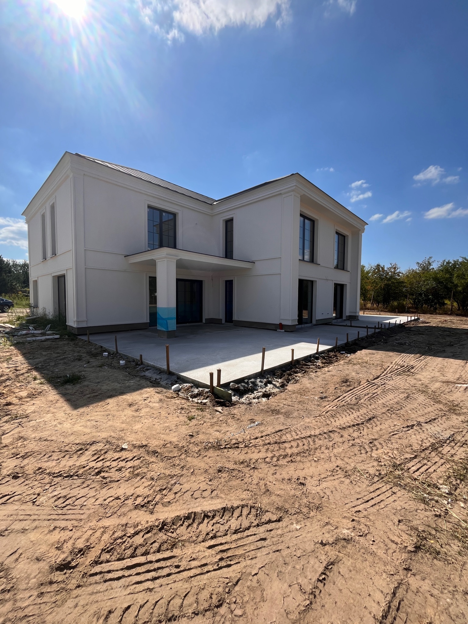 Dwukondygnacyjny biały budynek z płaskim dachem w trakcie budowy, widoczne fundamenty i wylewka tarasu, teren wokół z licznymi śladami opon.