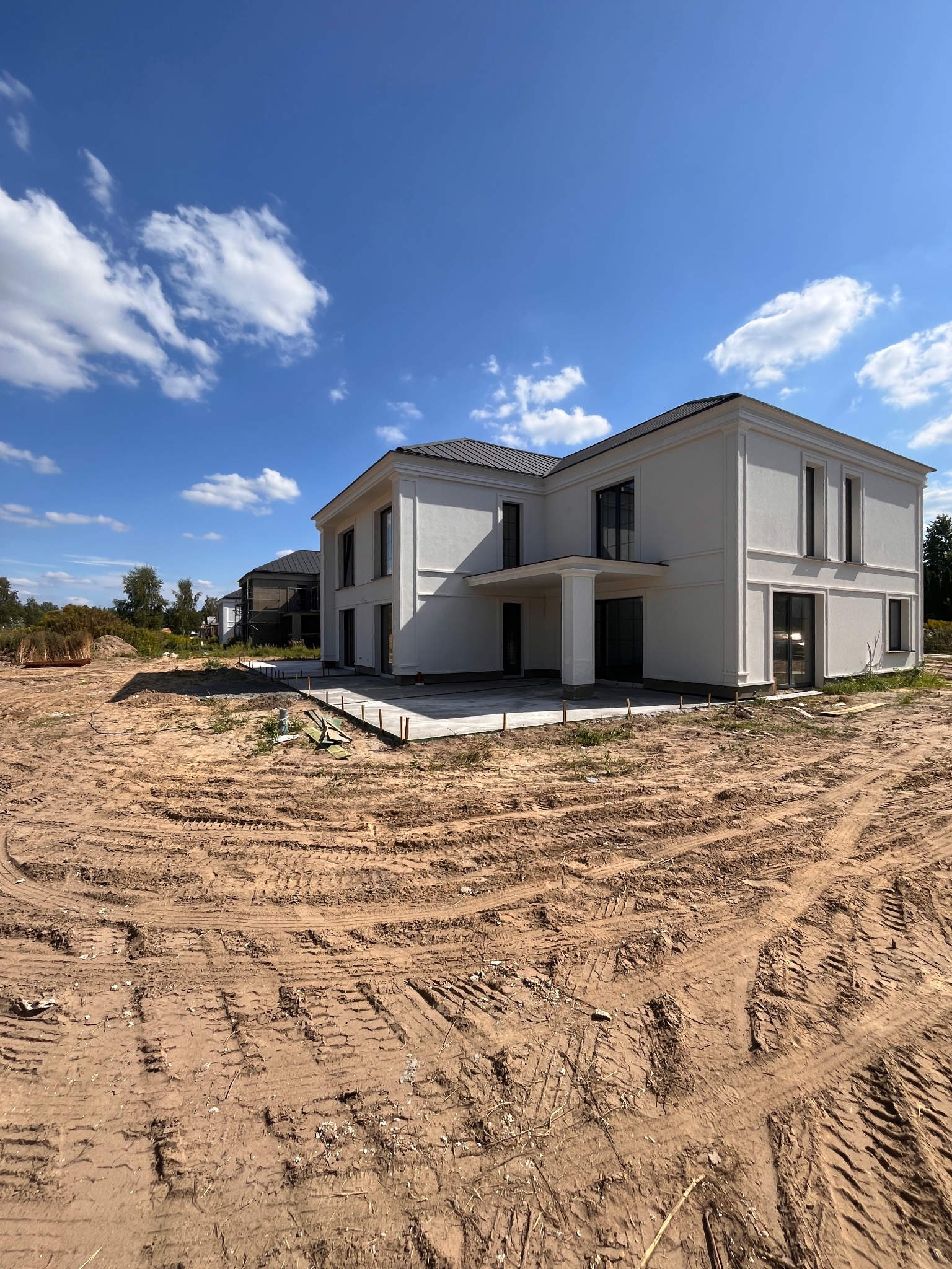 Dwukondygnacyjny budynek z białymi ścianami i ciemnym dachem w trakcie budowy, widoczne ślady opon na ziemi, błękitne niebo z chmurami w tle.