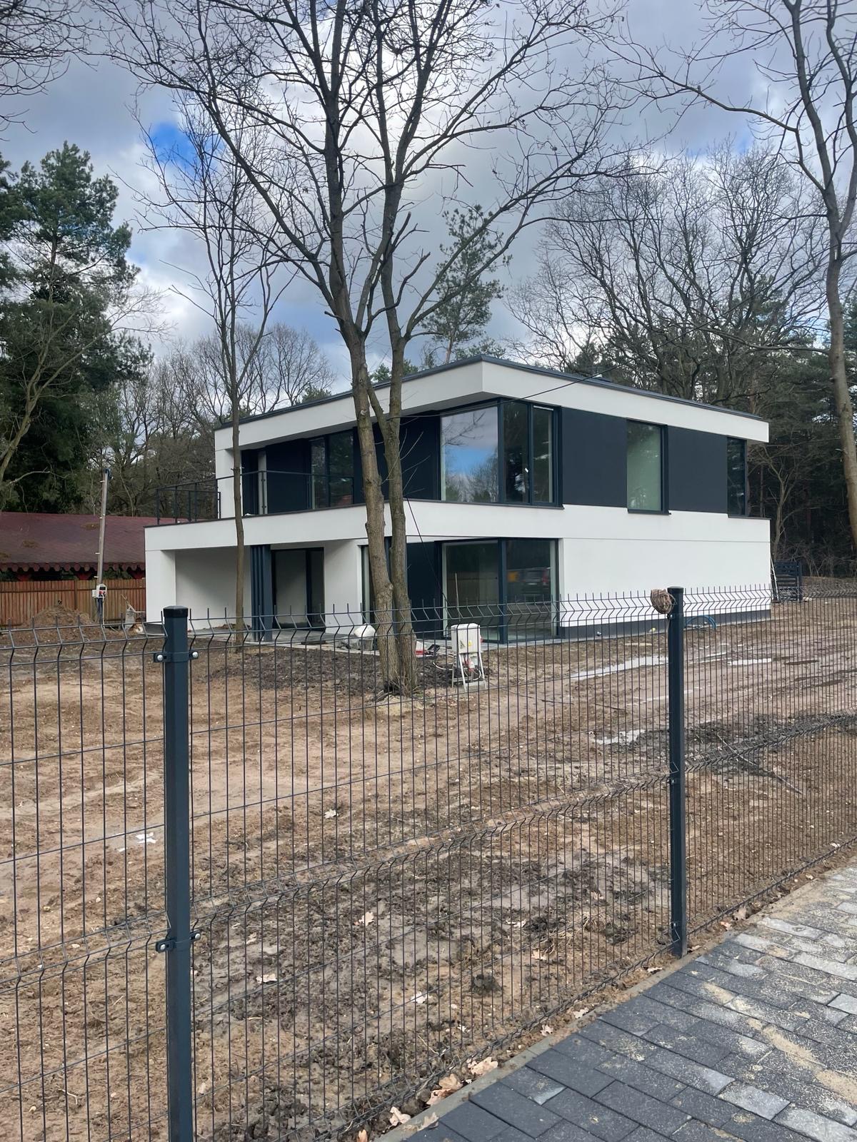 Nowoczesny, dwukondygnacyjny dom z płaskim dachem i dużymi oknami, otoczony drzewami i ogrodzeniem z siatki. Widoczne przygotowane podłoże wokół budynku.