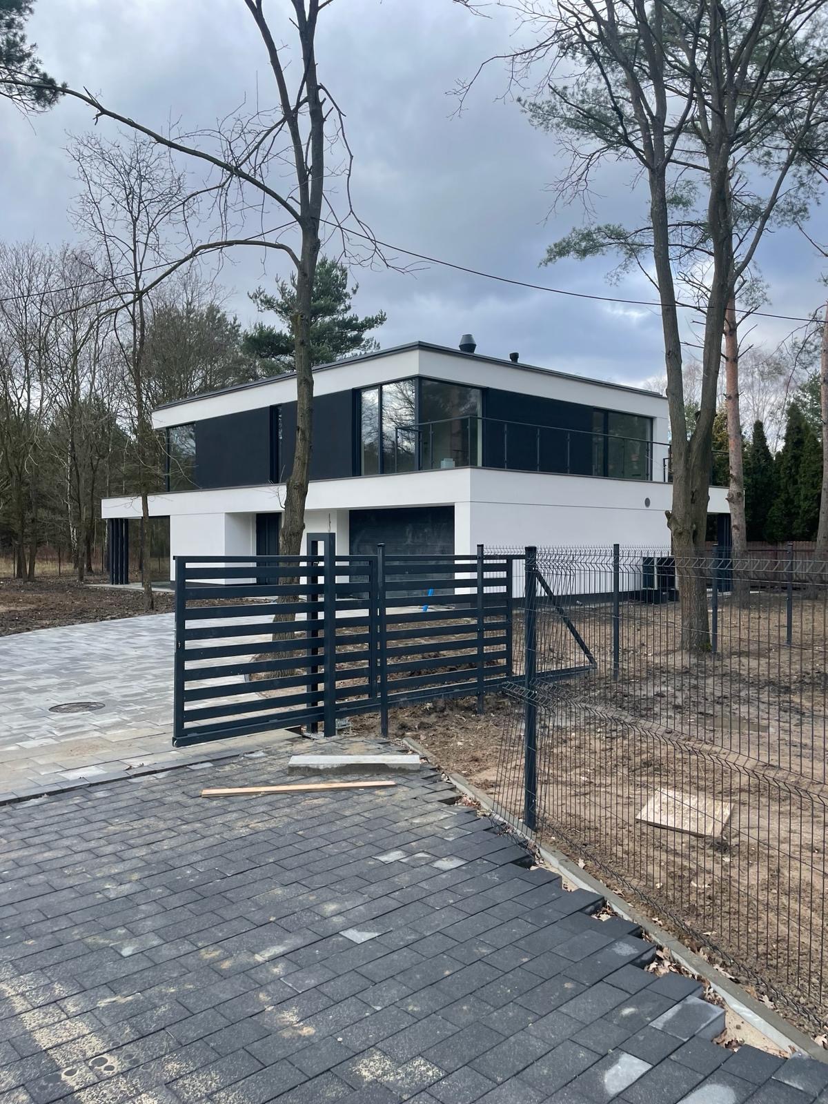Nowoczesny, dwukondygnacyjny dom z płaskim dachem, czarnymi akcentami i dużymi oknami, widziany zza czarnego ogrodzenia panelowego na nierównym podjeździe z kostki brukowej i ziemi.
