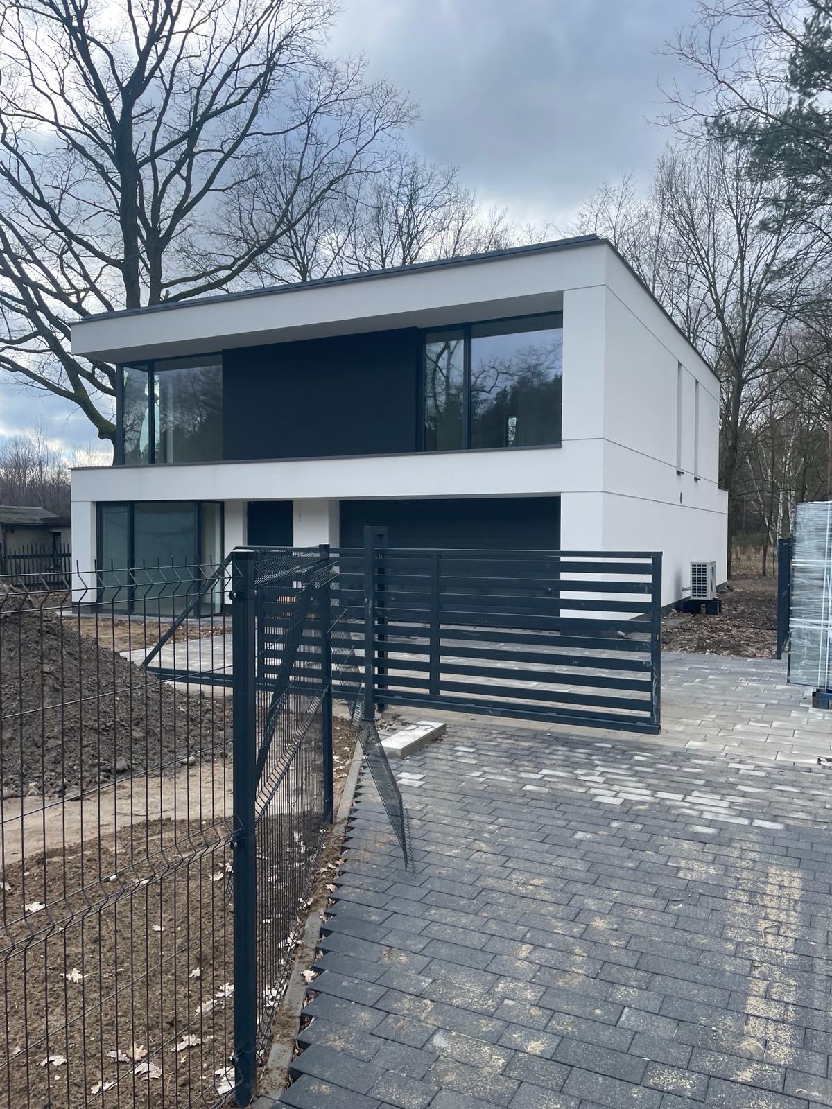 Nowoczesny, dwukondygnacyjny dom z płaskim dachem i dużymi oknami w Warszawie, widziany zza metalowego ogrodzenia i bramy wjazdowej. Ścieżka z kostki brukowej prowadzi do garażu. Na drugim planie...