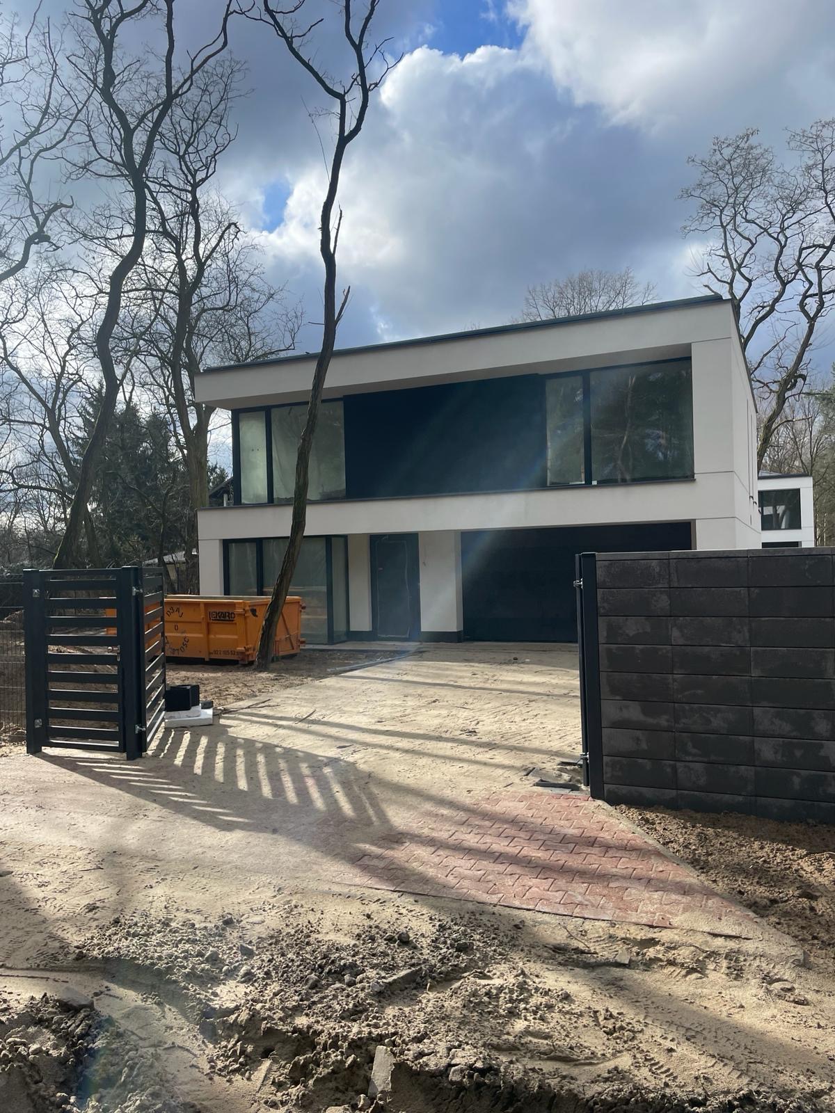 Nowoczesny, dwukondygnacyjny dom w trakcie budowy, z dużymi oknami i płaskim dachem, widoczny pomarańczowy kontener na gruz i fragment ogrodzenia.