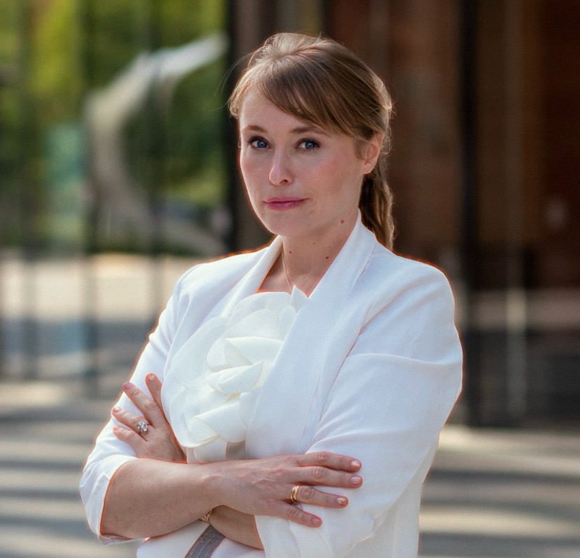 Portret kobiety w białym żakiecie z ozdobną różą, skrzyżowane ręce, pierścionki, Warszawa, budynek w tle.