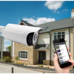 Kamera IP z anteną na tle domu, obsługiwana przez smartfon. Aplikacja monitoringu z podglądem na żywo z różnych pomieszczeń.