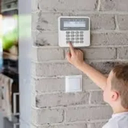 Dziecko obsługuje panel alarmowy na ceglanej ścianie. Widoczny biały panel z klawiaturą numeryczną i przyciskami funkcyjnymi. Nowoczesny system alarmowy w domu.