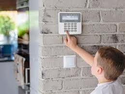 Dziecko obsługuje panel alarmowy na ceglanej ścianie. Widoczny biały panel z klawiaturą numeryczną i przyciskami funkcyjnymi. Nowoczesny system alarmowy w domu.