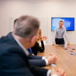 Prelegent w jasnej koszuli prezentuje materiał na dużym ekranie podczas spotkania w sali konferencyjnej, uczestnicy siedzą przy długim drewnianym stole.