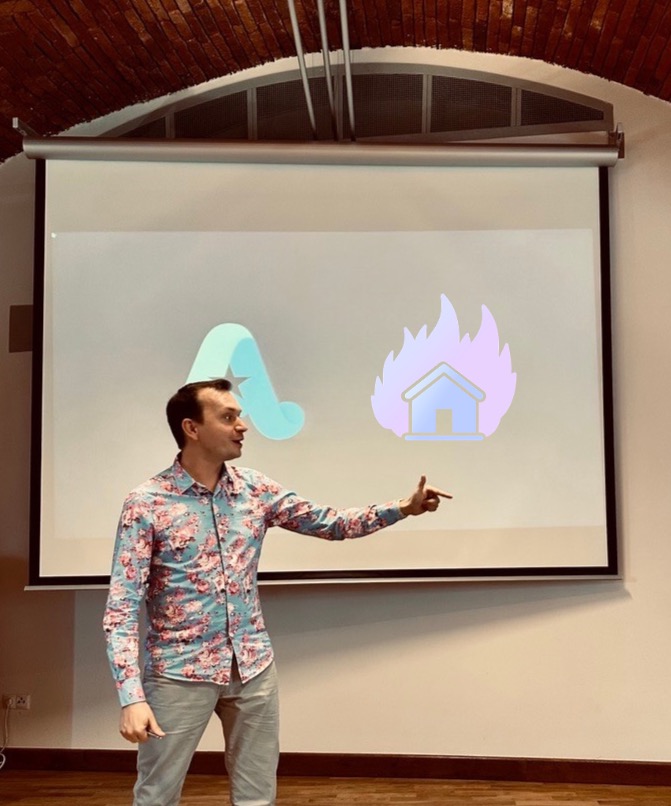 Prelegent w kwiecistej koszuli prezentuje slajd z logo firmy i płonącym domem na szkoleniu biznesowym. Sala konferencyjna z ceglanym sklepieniem.