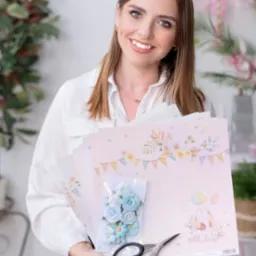 Uśmiechnięta kobieta trzyma w dłoniach arkusze papieru z motywem dziecięcym, paczkę błękitnych kwiatków i nożyczki, w tle dekoracje kwiatowe.