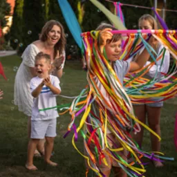 Radosne dzieci na zielonej trawie, w tle kobieta klaszcze. Chłopiec trzyma kolorową obręcz z wstążkami, a w powietrzu unoszą się bańki mydlane. Uśmiechy i dobra zabawa.