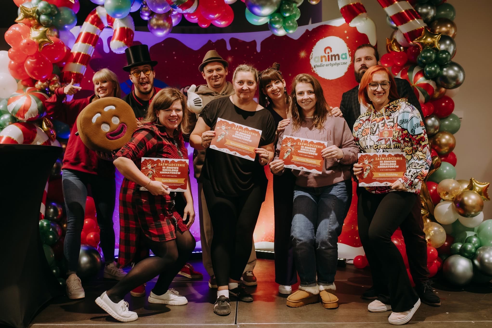 Grupa osób pozuje z uśmiechem, trzymając certyfikaty ukończenia zimowego szkolenia dla animatorów, na tle dekoracji z balonów i pluszowego piernika.