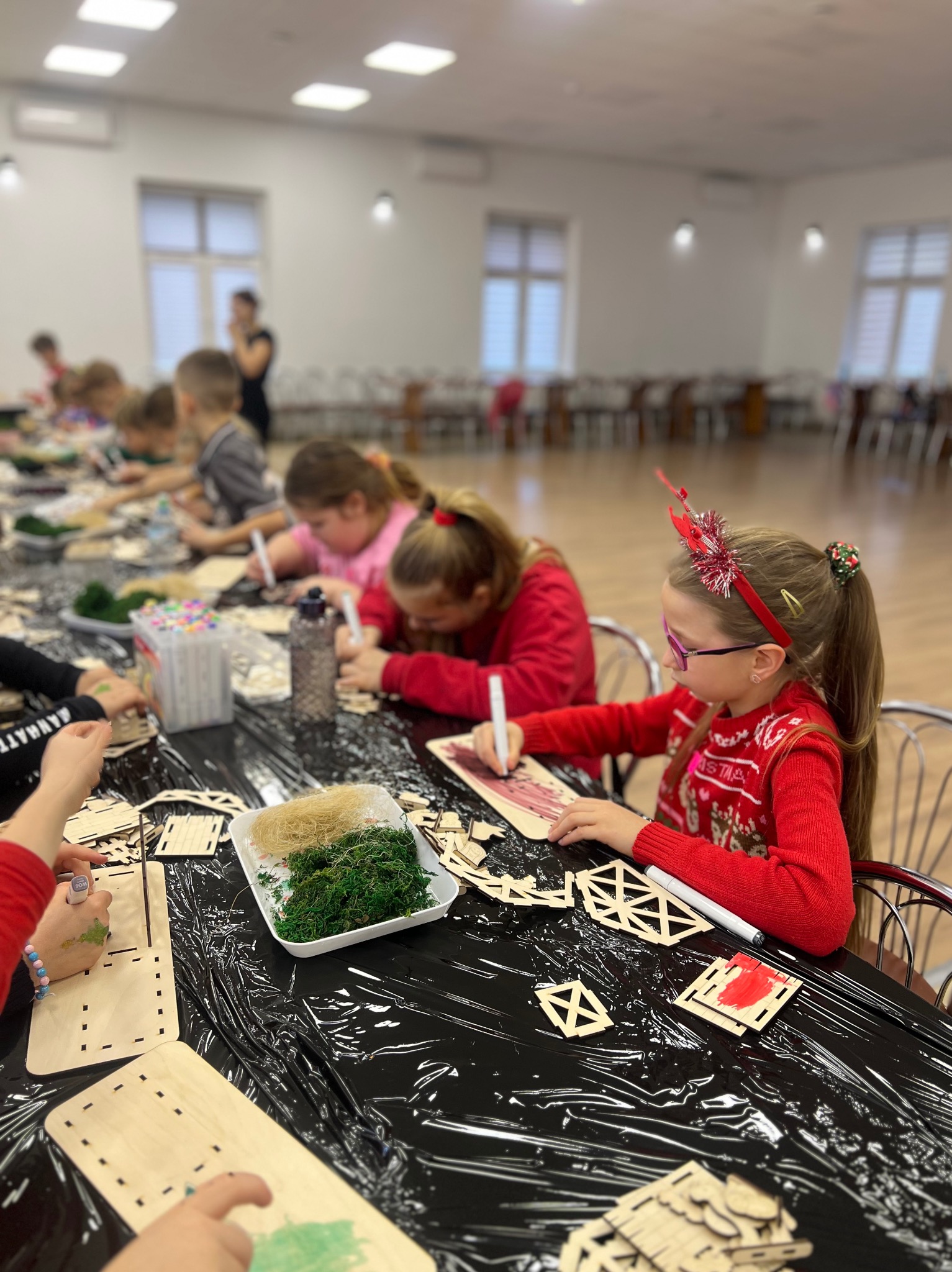 Grupa dzieci przy stole podczas warsztatów plastycznych, malowanie drewnianych elementów dekoracyjnych na czarnym obrusie, w tle widoczne okna i osoba dorosła.