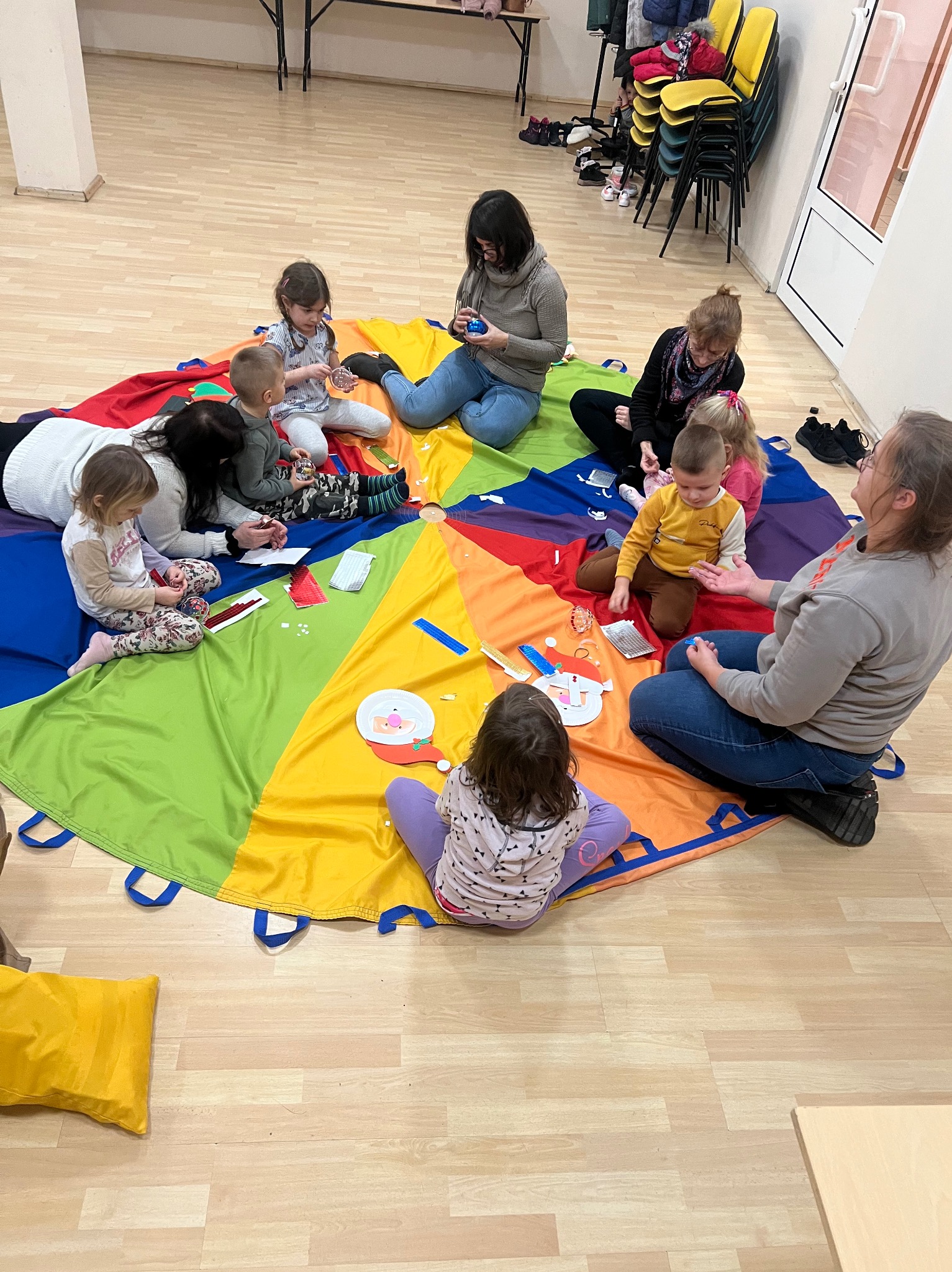 Grupa dzieci i dorosłych siedzi na podłodze na kolorowej chuście, tworząc ozdoby świąteczne z papieru i innych materiałów.