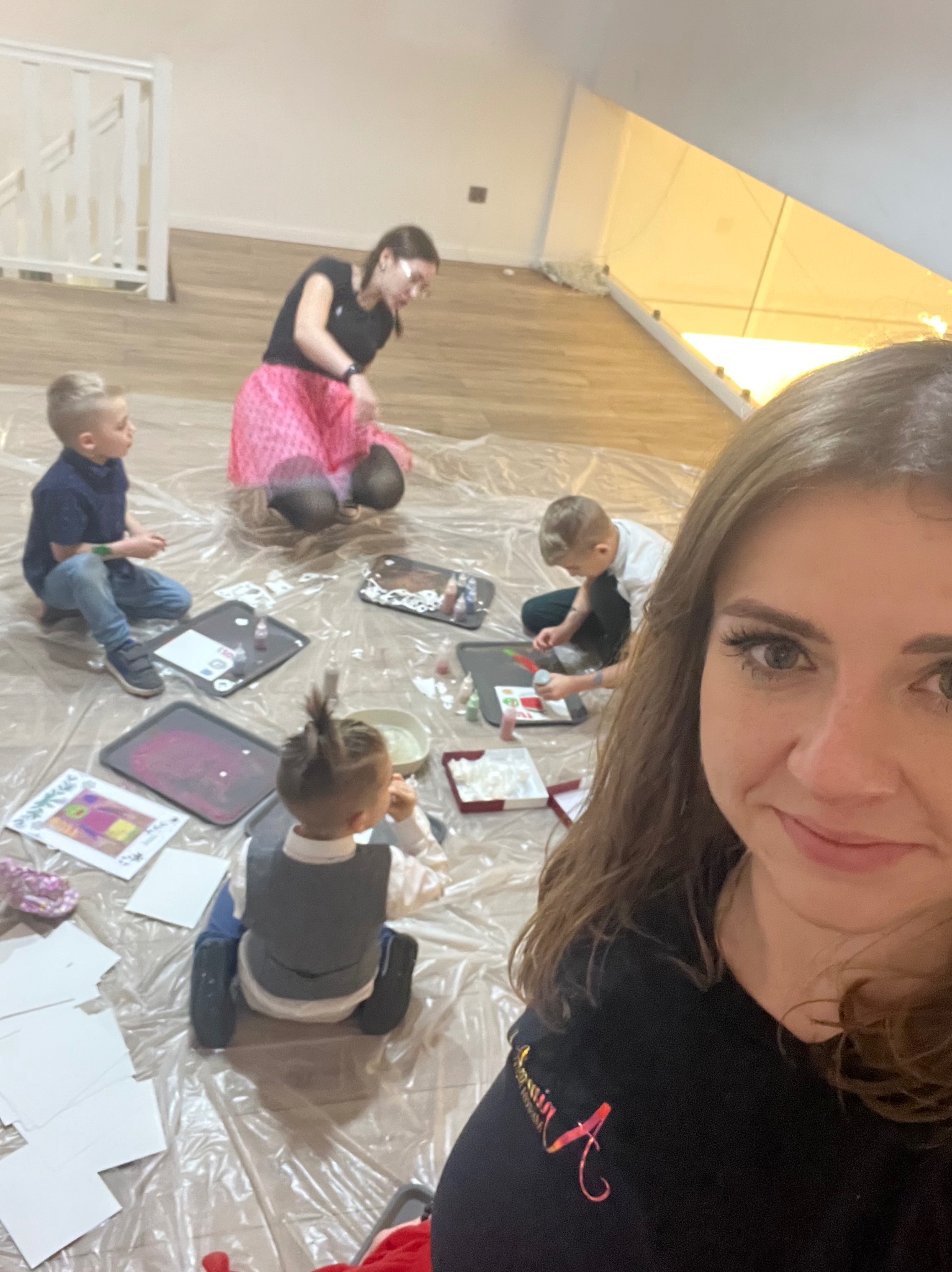 Kobieta robiąca selfie podczas warsztatów plastycznych dla dzieci, na podłodze rozłożona folia ochronna, dzieci malują farbami, widoczne prace plastyczne i materiały do malowania.
