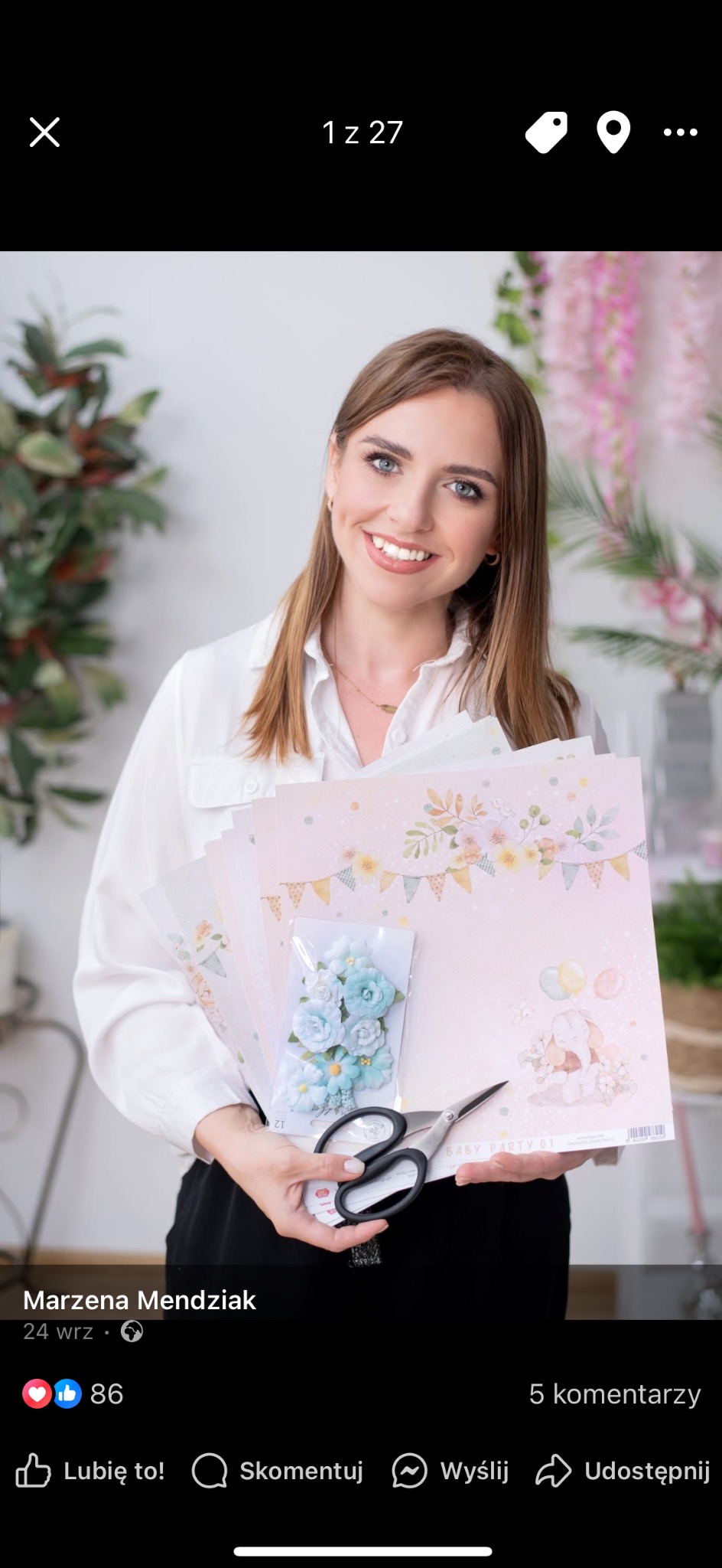 Uśmiechnięta kobieta trzyma w dłoniach arkusze papieru z motywem dziecięcym, paczkę błękitnych kwiatków i nożyczki, w tle dekoracje kwiatowe.