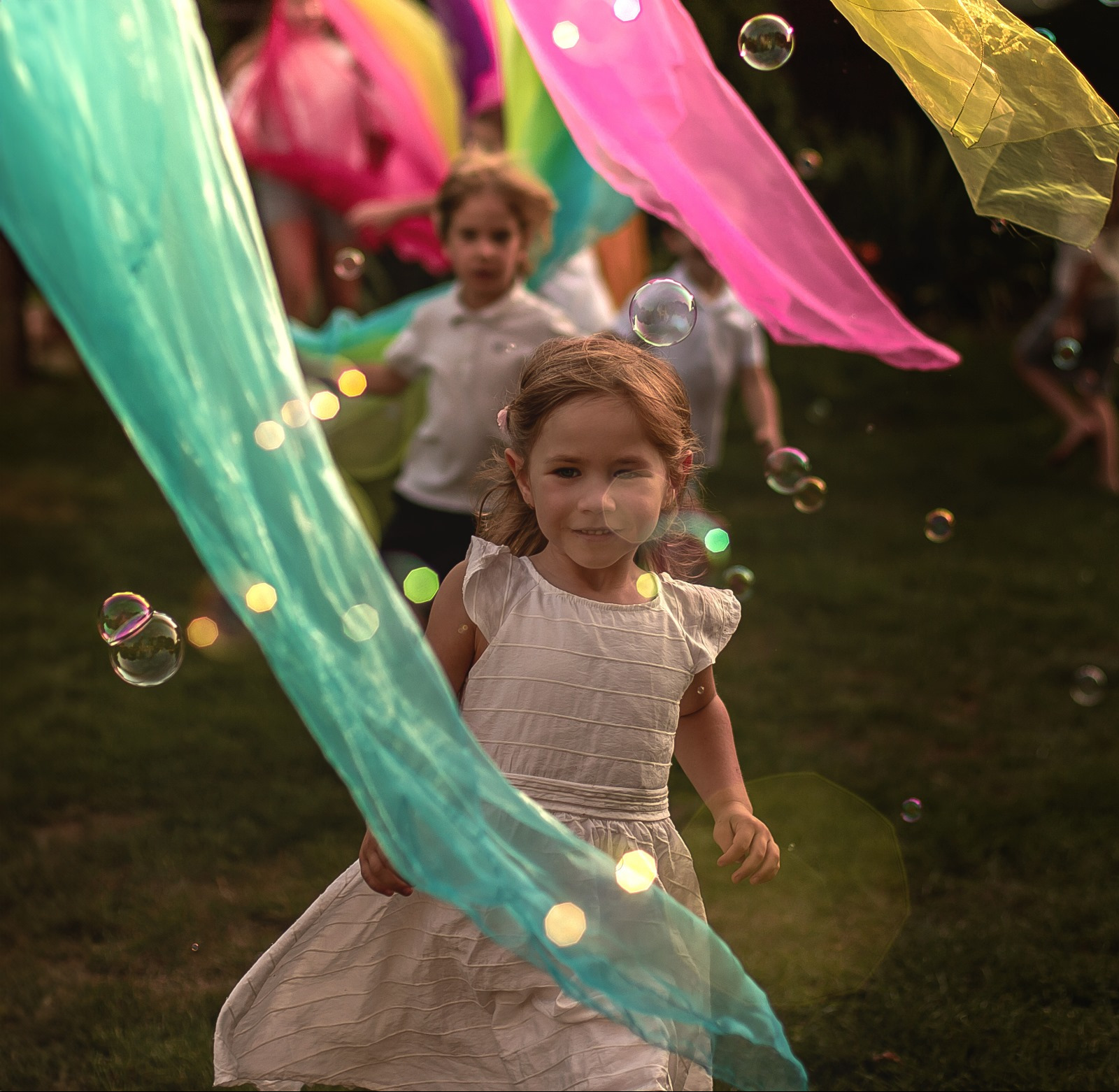Uśmiechnięta dziewczynka w białej sukience bawi się na trawie z kolorowymi chustami i bańkami mydlanymi, w tle inne dzieci i zieleń.