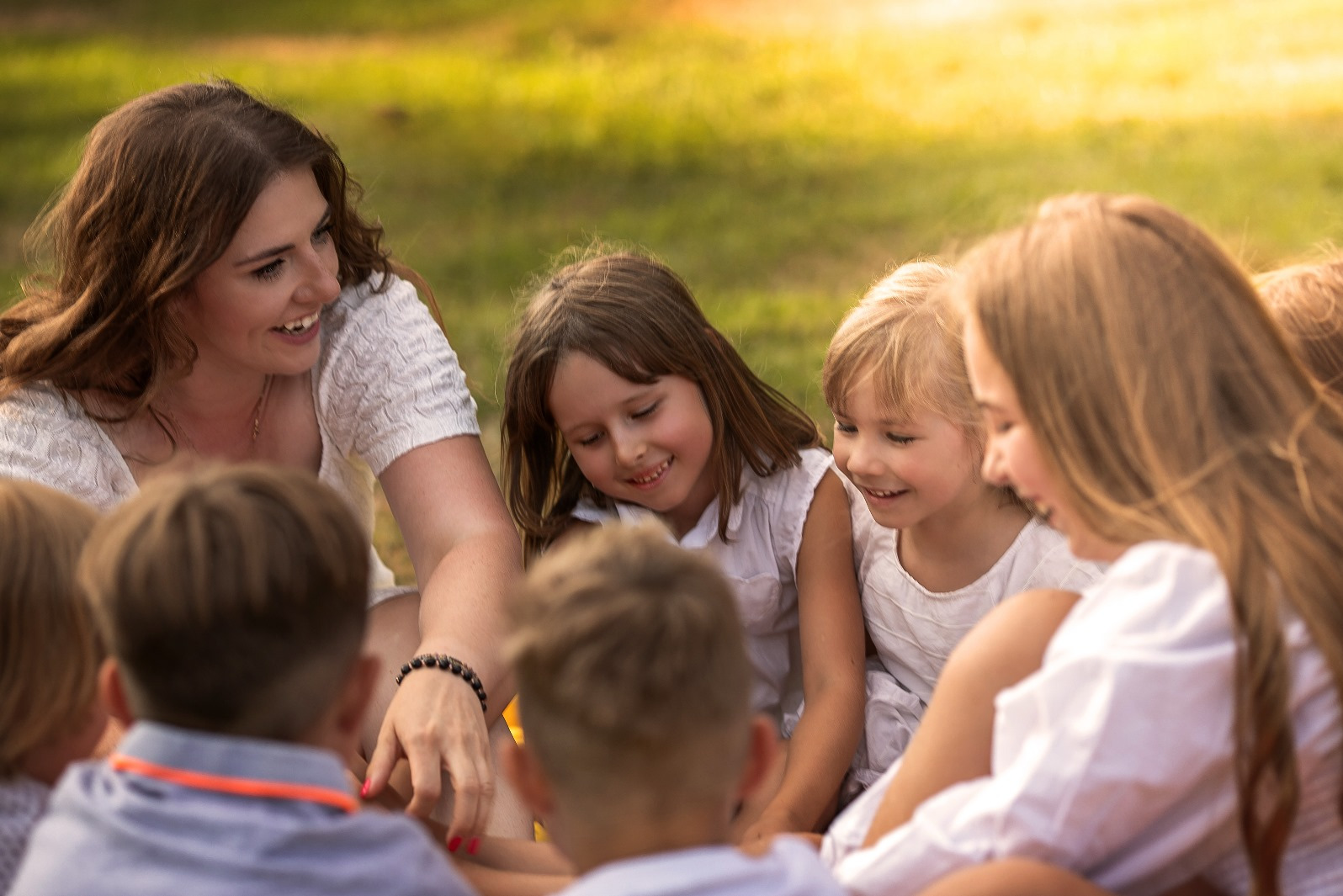 Uśmiechnięta animatorka w otoczeniu dzieci na trawie, w słoneczny dzień. Grupa siedzi w kręgu, skupiona na wspólnej zabawie, w tle zielona trawa.