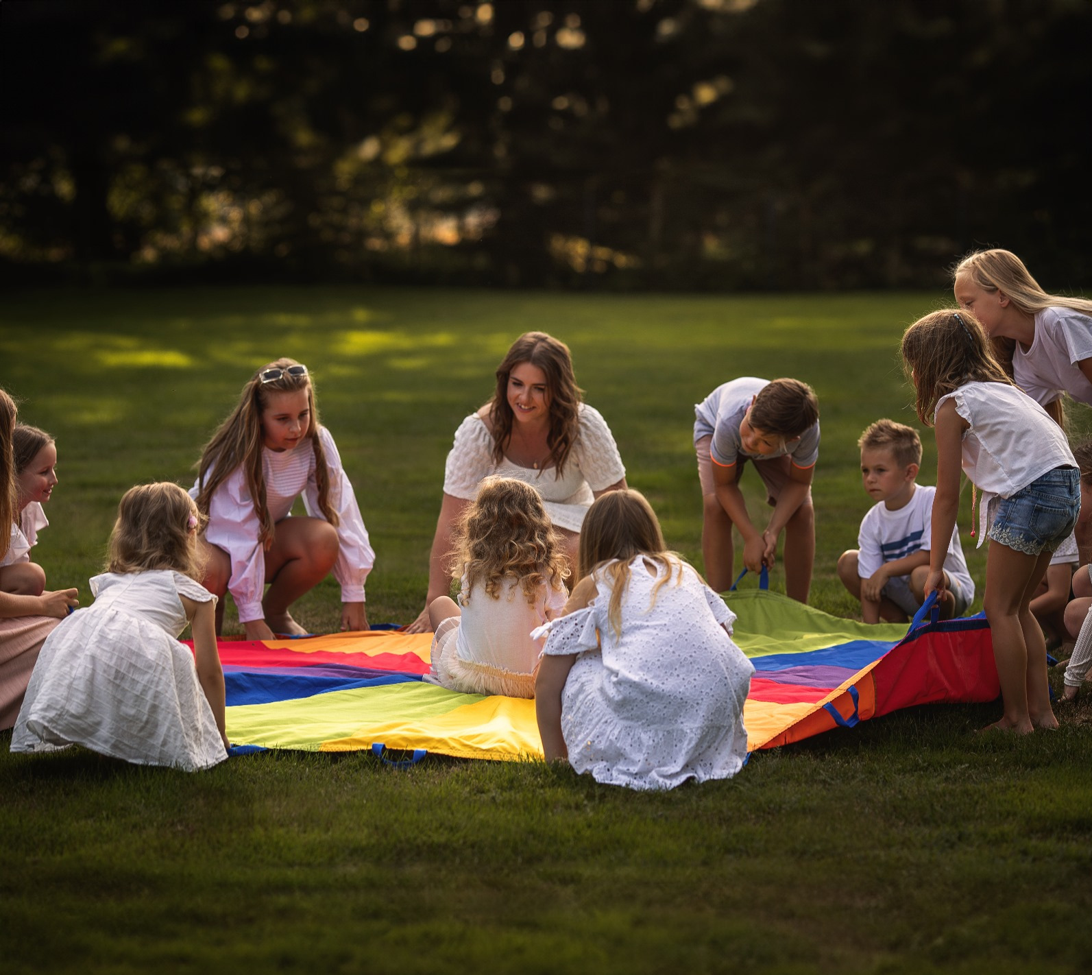 Grupa dzieci i animatorka bawi się kolorową chustą animacyjną na trawie. Uśmiechnięta kobieta w białej sukience siedzi pośród dzieci. Zabawa na świeżym powietrzu.