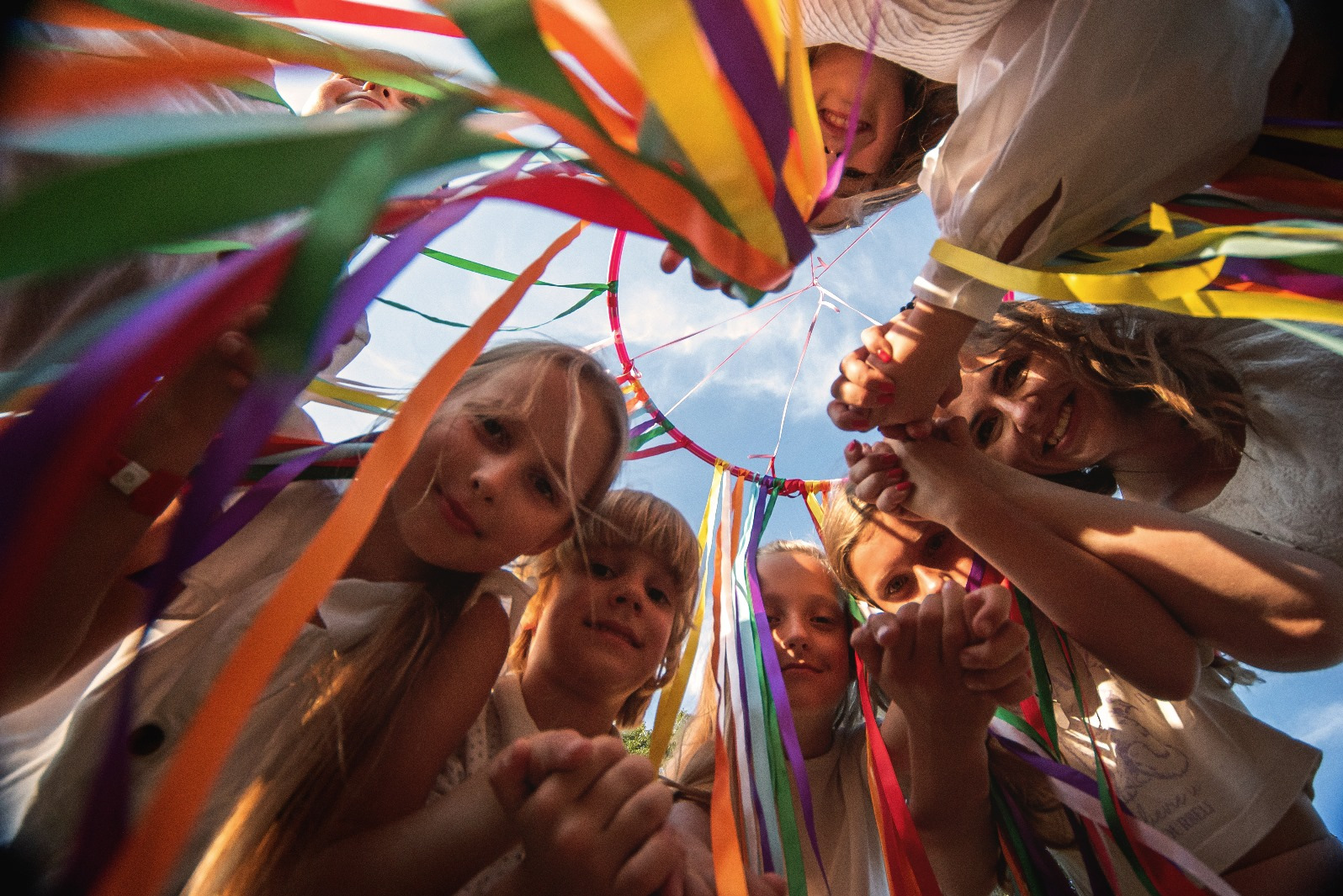 Grupa uśmiechniętych dzieci i dorosłych spogląda w dół, otoczona kolorowymi wstążkami na tle błękitnego nieba, tworząc radosną i pełną energii kompozycję.