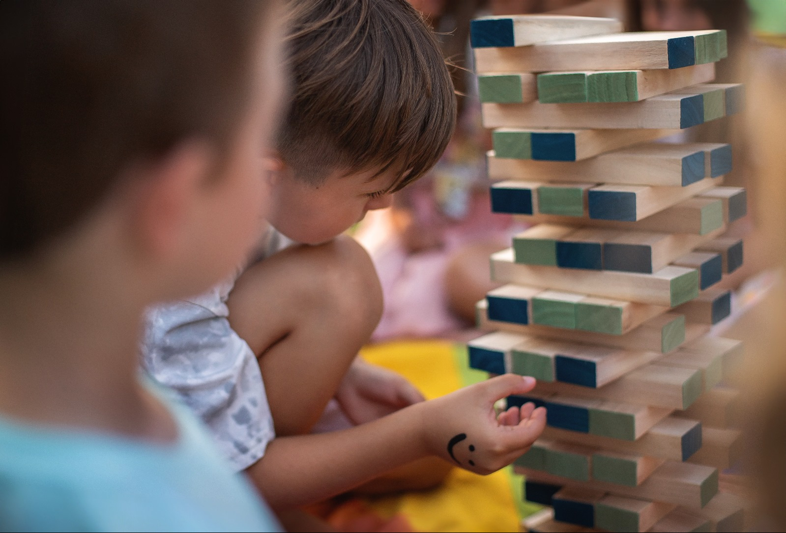 Dziecko z uśmiechem na ręce gra w drewnianą wieżę Jenga z kolorowymi elementami. Widoczne skupienie i interakcja podczas zabawy grupowej.